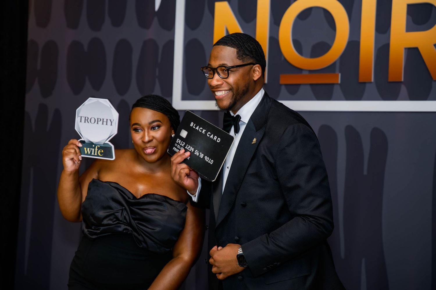 A glamorous couple poses at a formal event against a backdrop with 'NOIR' signage.