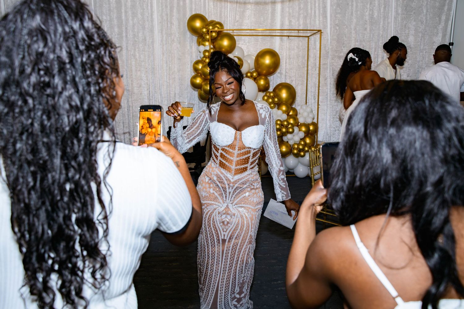 Guests taking photos at an elegant party with gold balloon decorations and white decor.
