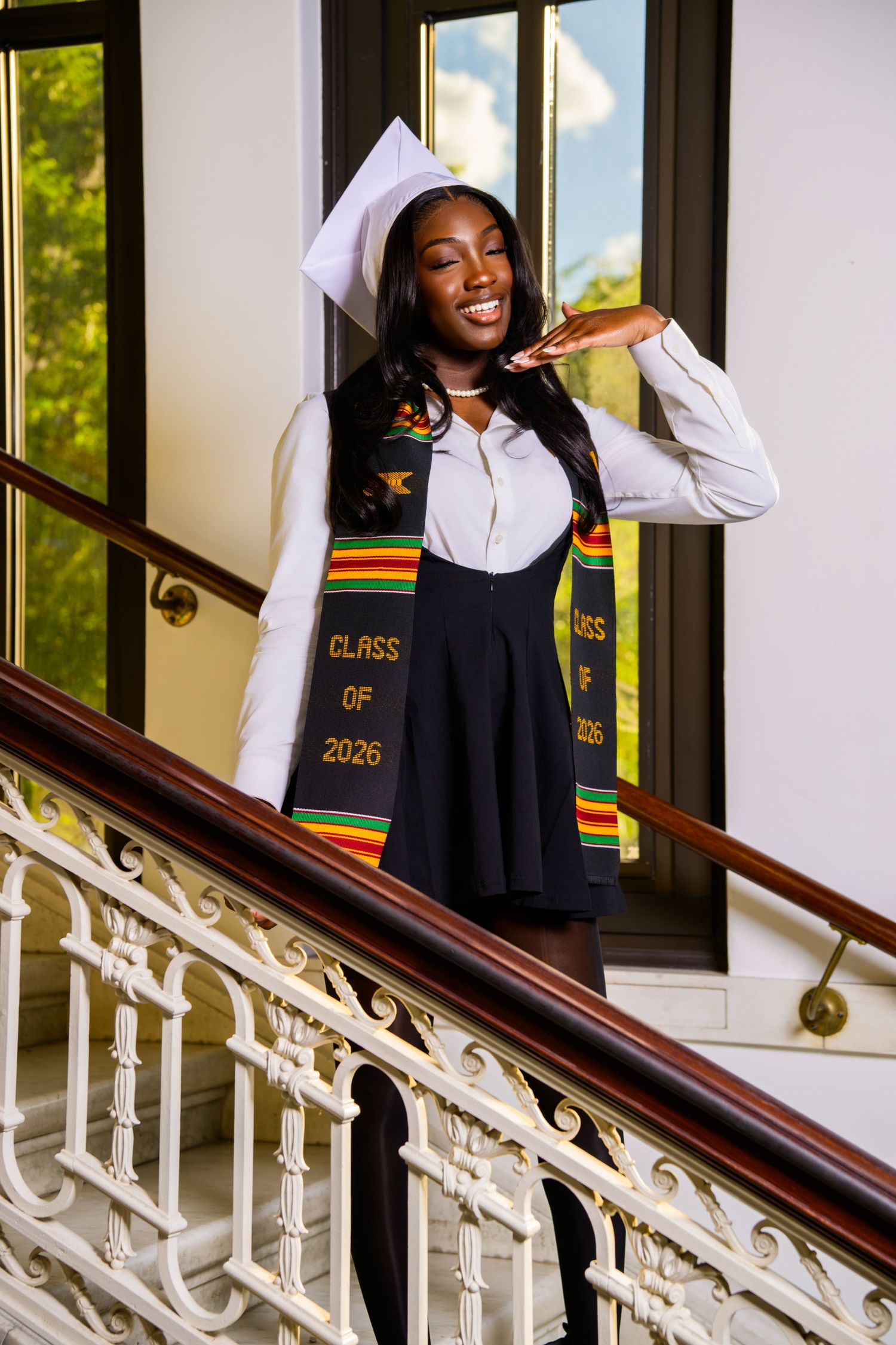 Smiling Black woman in graduation cap and Class of 2026 kente stole poses on elegant staircase in white blouse.