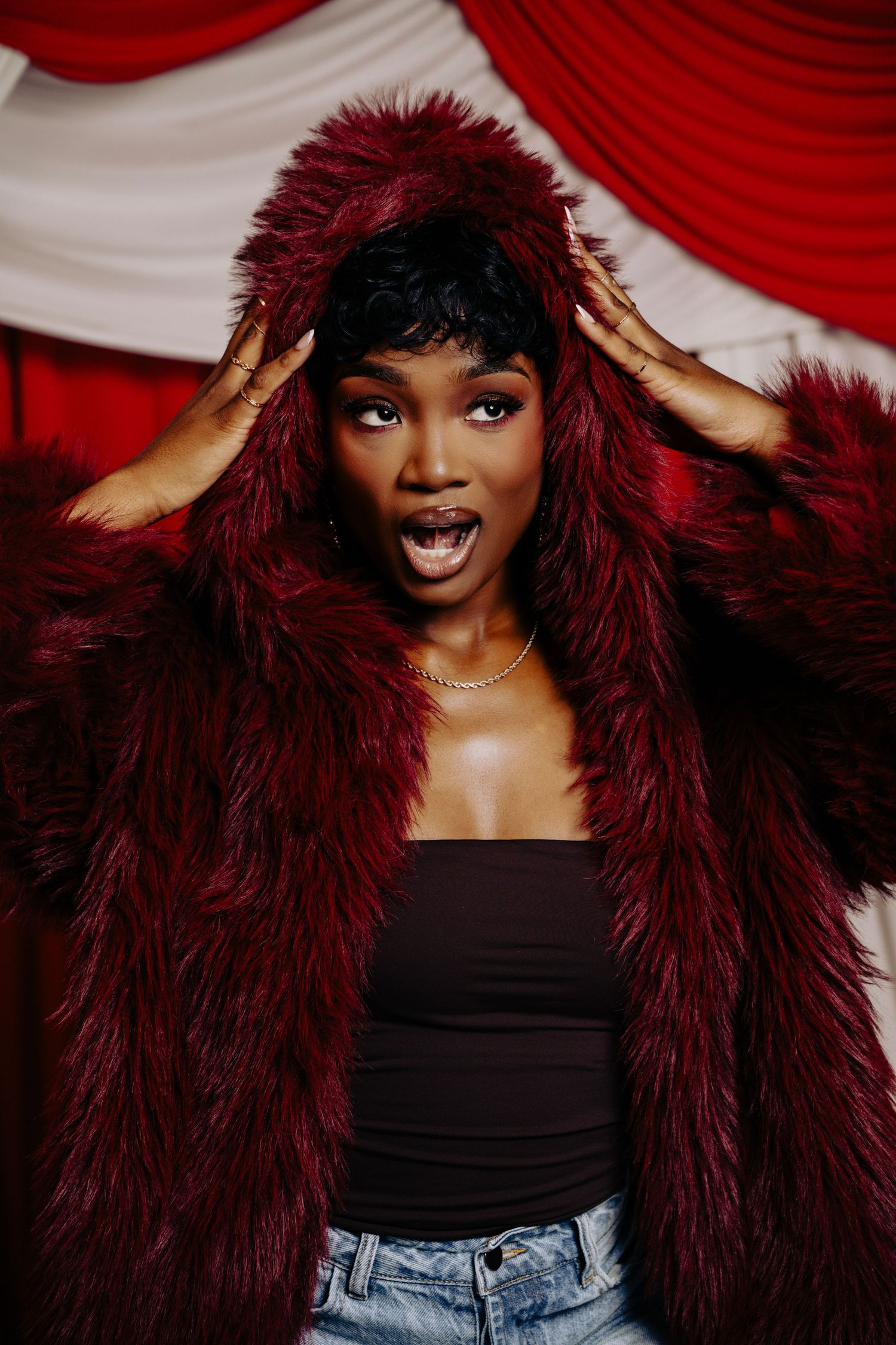 A bright smile radiates against a dramatic burgundy faux fur coat and black top with red curtain backdrop.