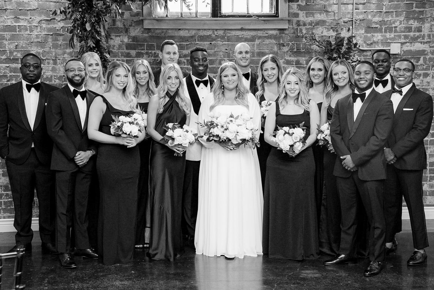 Wedding party lined up against brick wall for formal group photo.