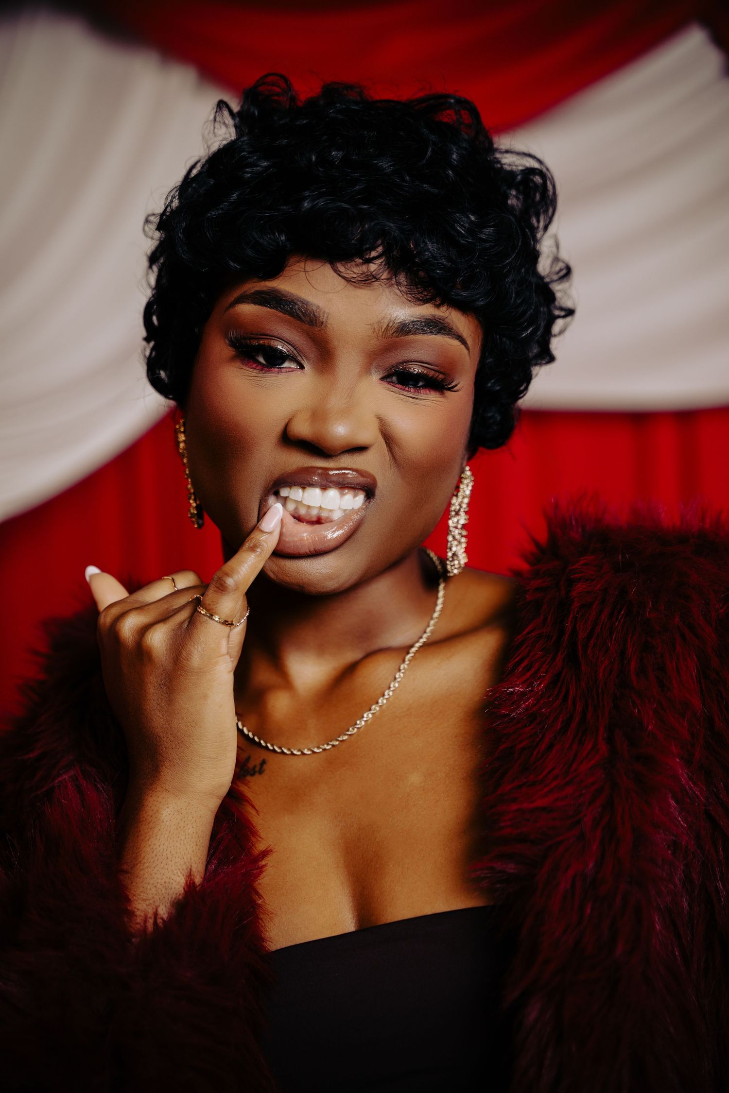Glamorous portrait against red and white backdrop featuring curly black hair, sparkly jewelry and burgundy feather boa.