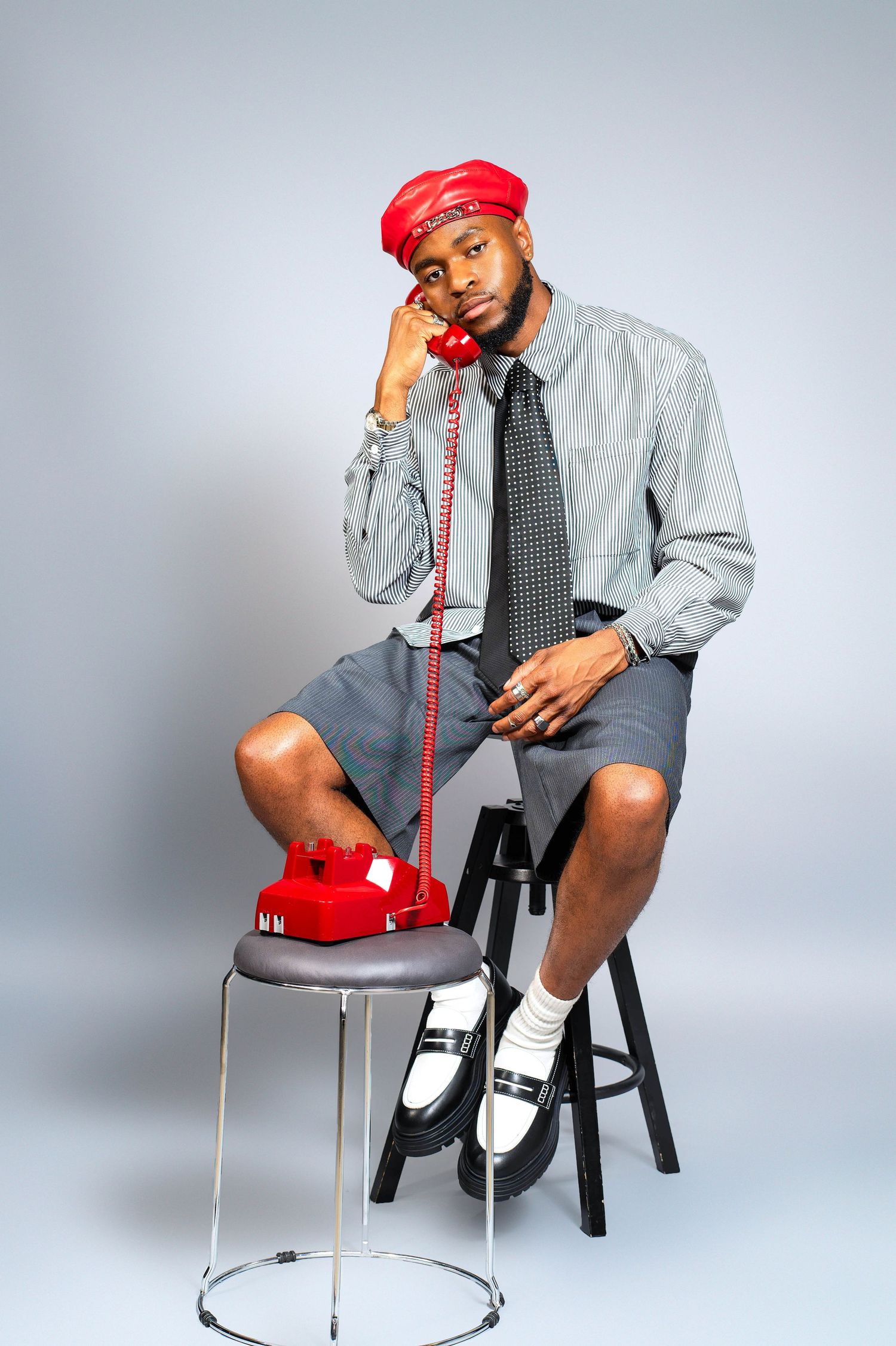 Person in business attire and red cap posing with vintage red telephone on stool.