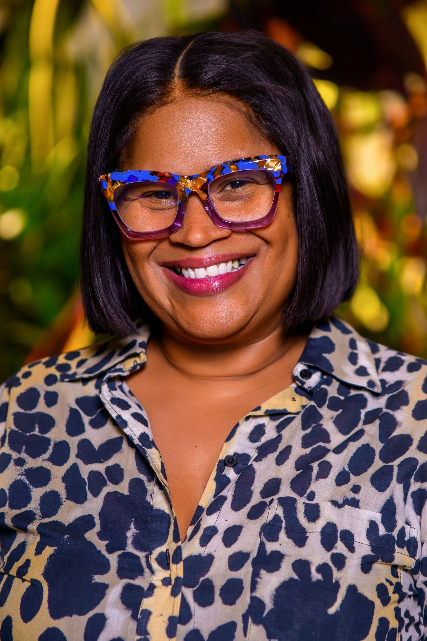 Smiling woman with blue tortoiseshell glasses and bob haircut wearing leopard print blouse outdoors.