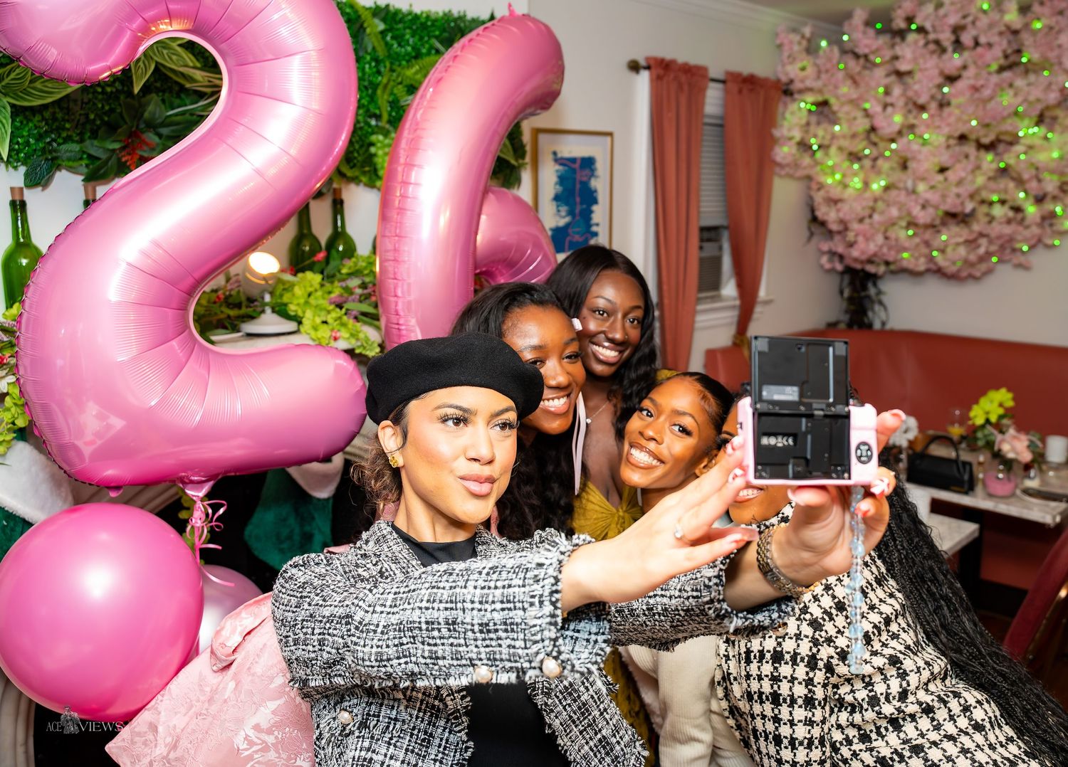 A group taking selfies together at a party with pink number balloons and floral wall decorations.
