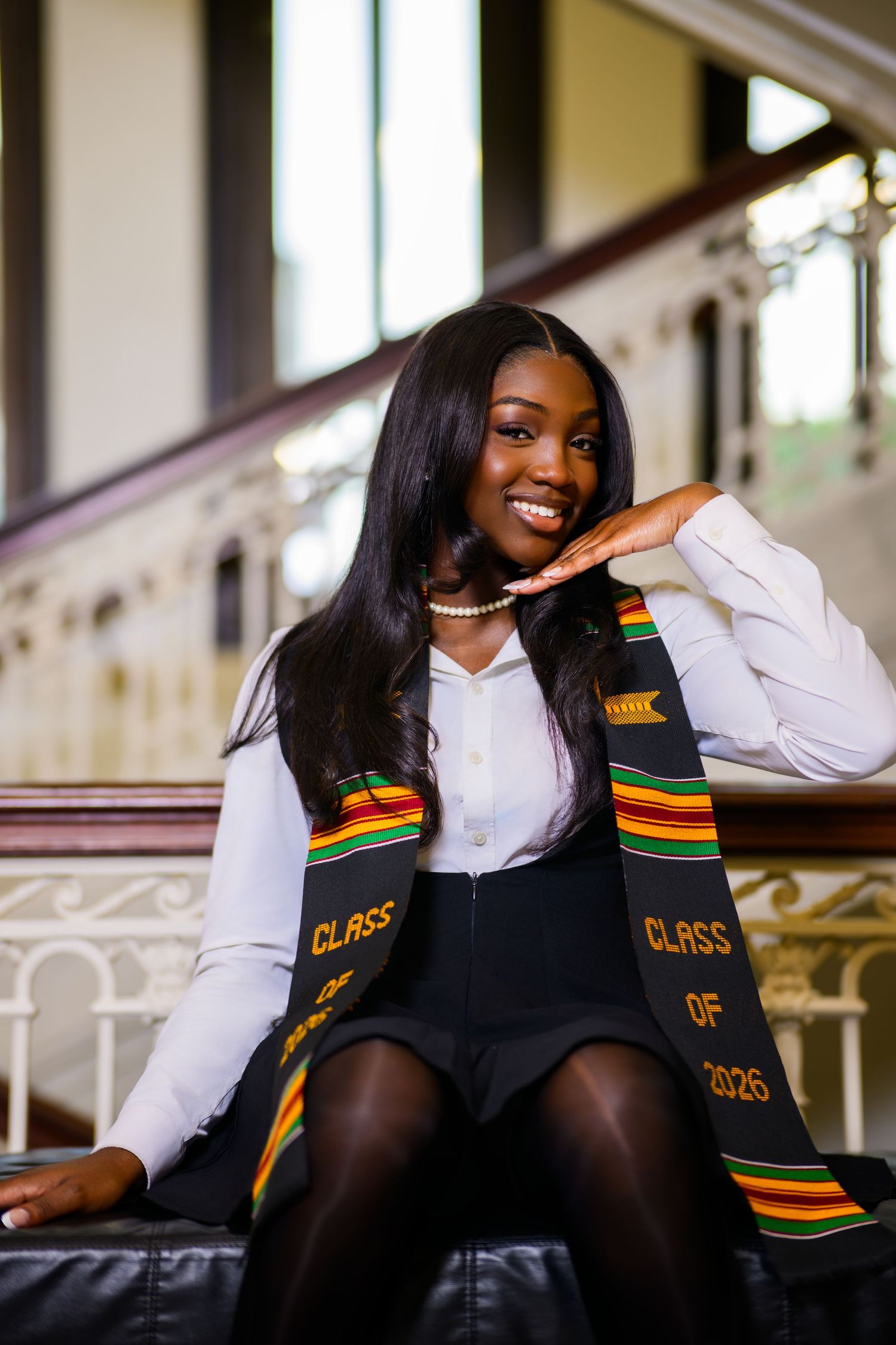 Smiling Black woman in white blouse and kente cloth Class of 2026 graduation stole seated on stairs.
