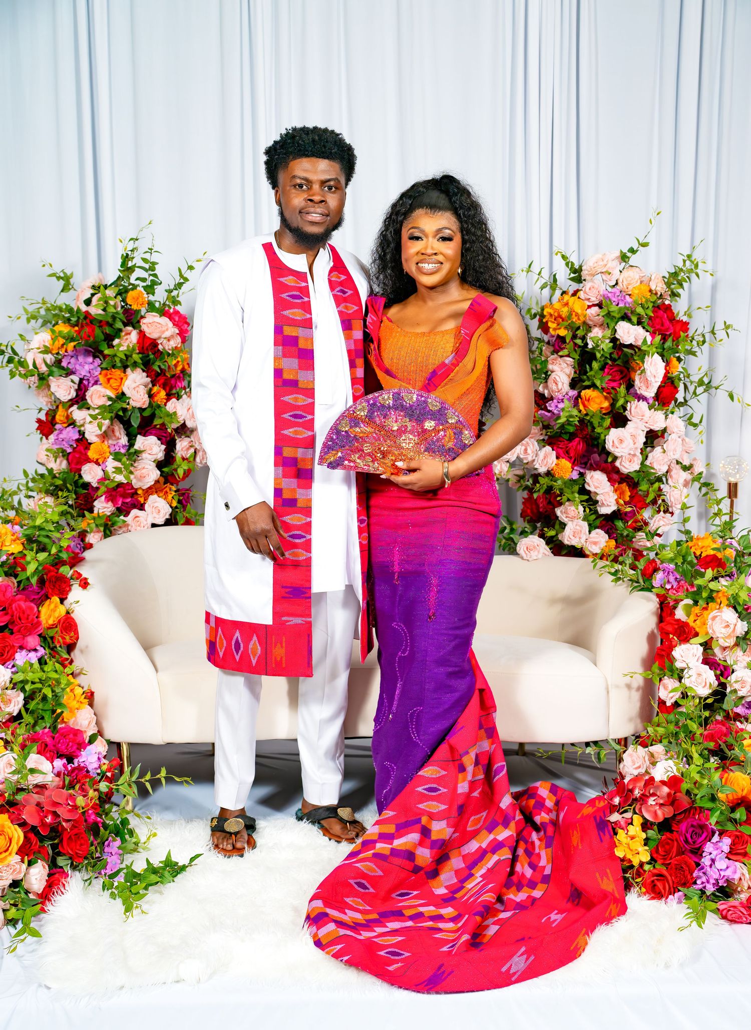 Couple in formal attire posing against white backdrop with vibrant floral arrangements.