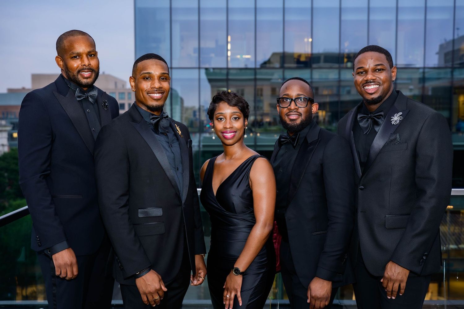 A group in formal attire poses together at an evening event with city views in the background.