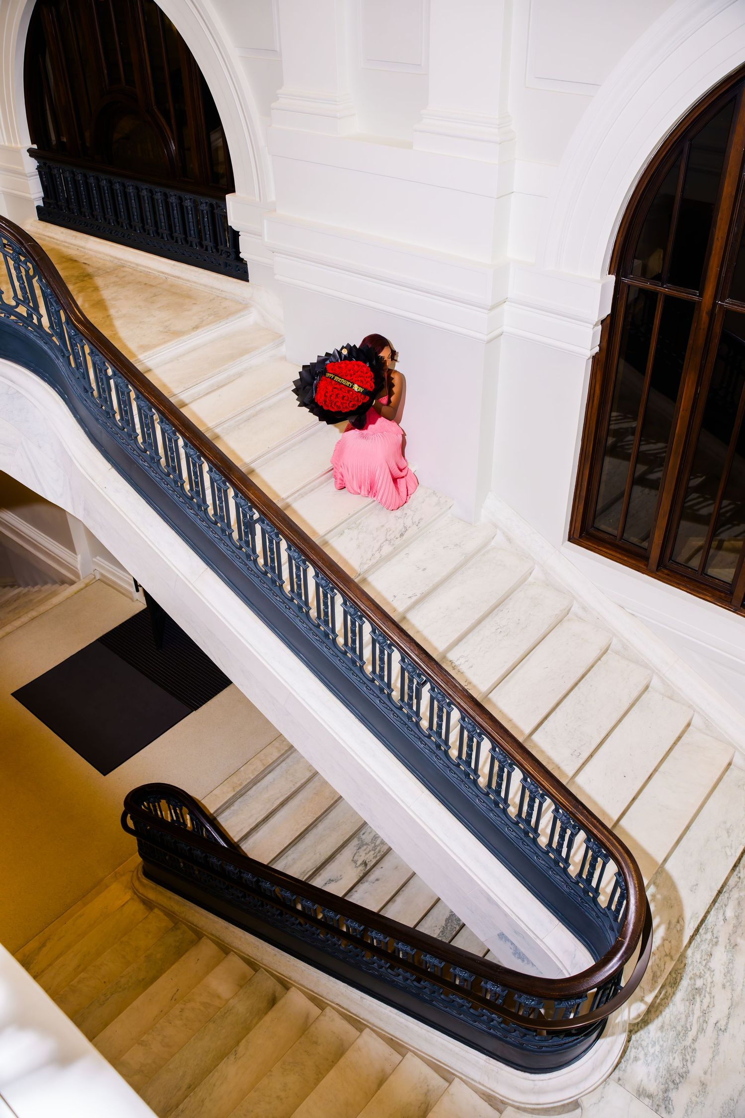 Woman in pink dress with red floral headpiece poses gracefully on elegant white marble staircase with dark railings.