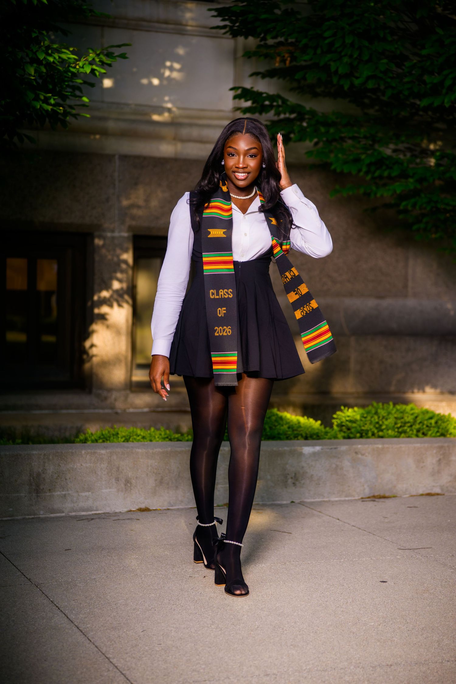 Young Black woman in Class of 2026 kente stole and black skirt posing outdoors near university building.