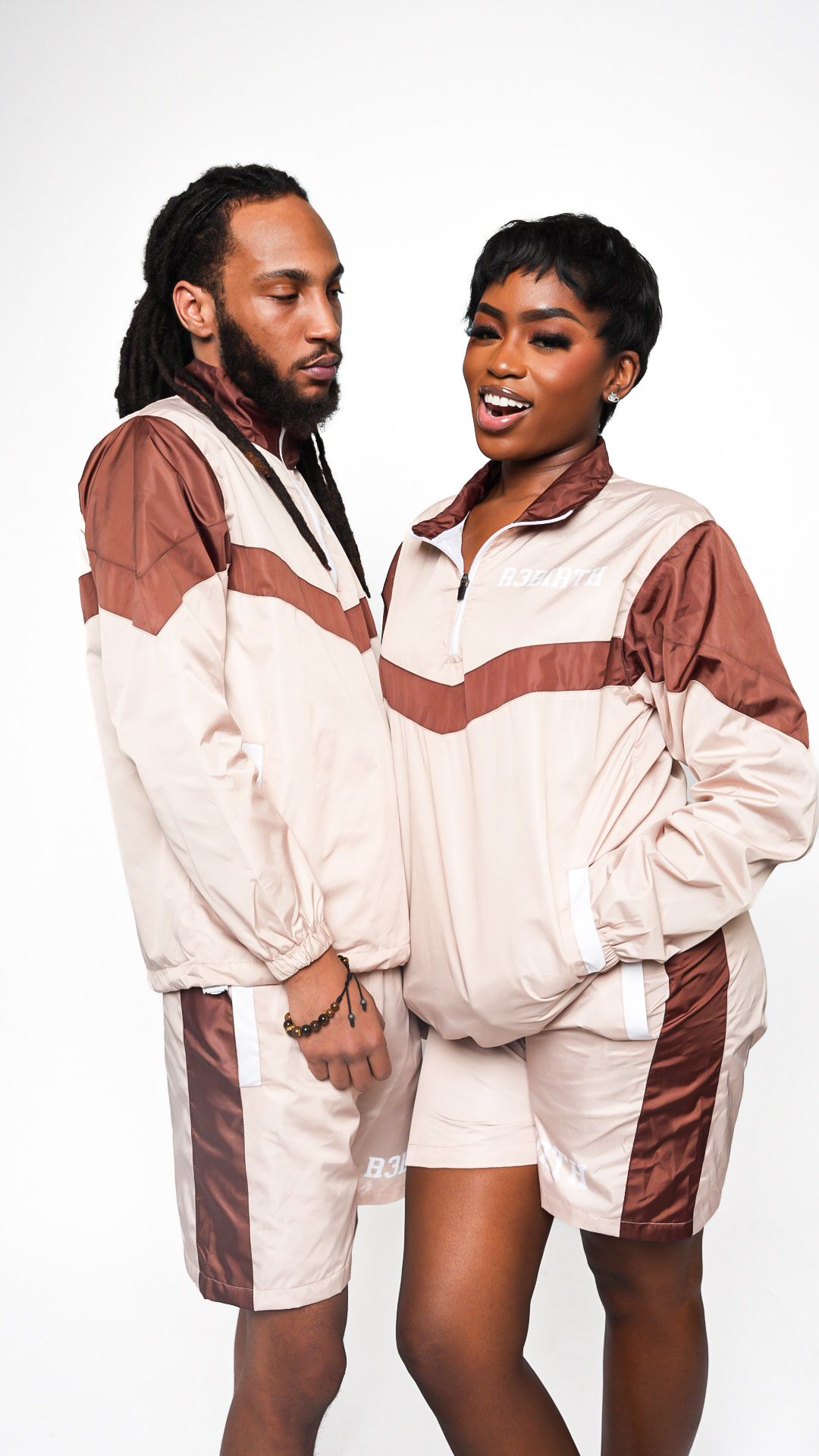 Fashion photoshoot with two models in cream and brown windbreaker jackets against white background.