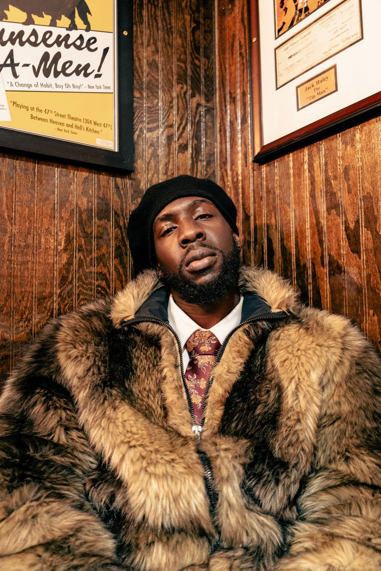 Person wearing luxurious fur coat and hat posing against wooden wall with framed artwork.