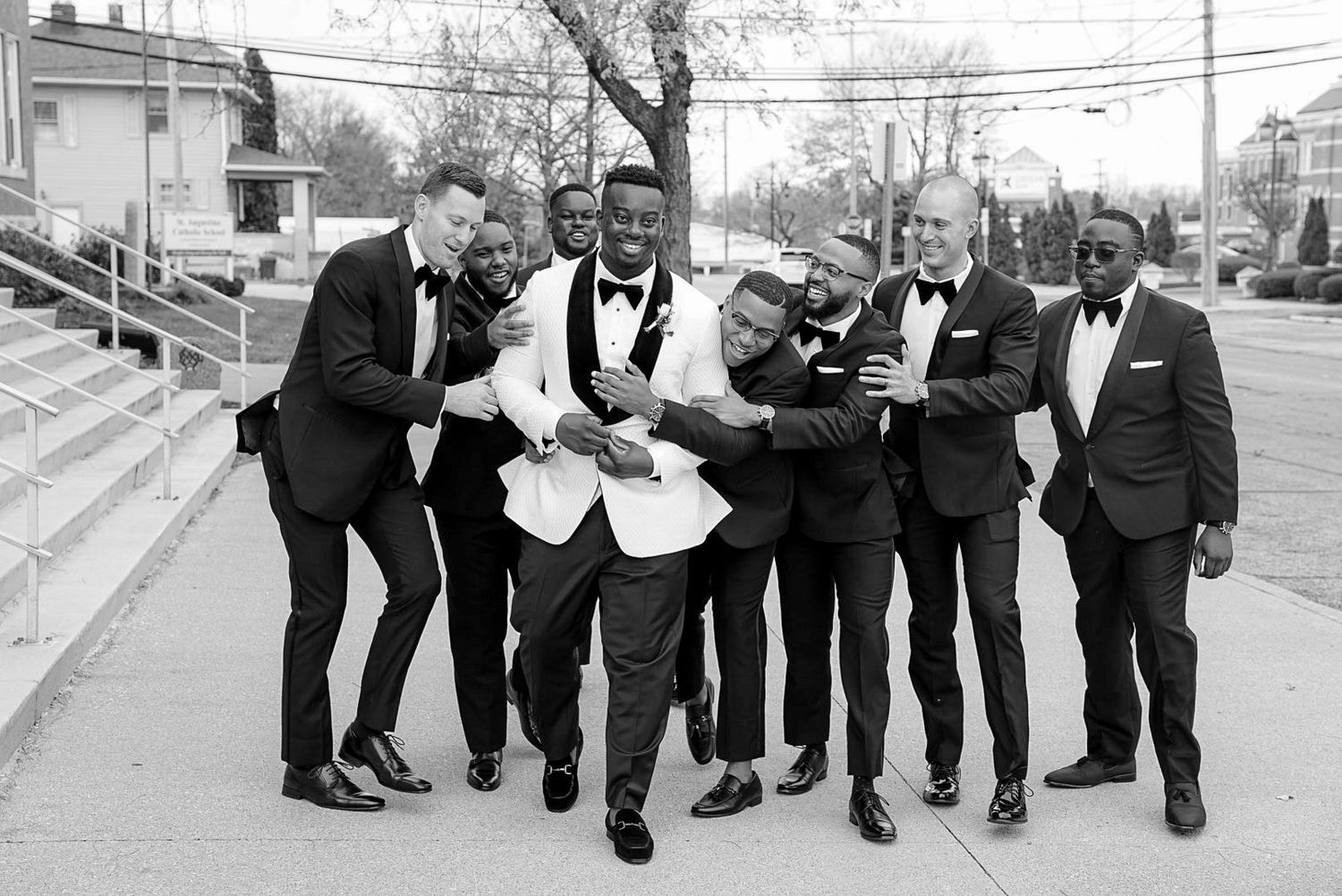 A group of groomsmen in black suits striking fun poses for a wedding photo.
