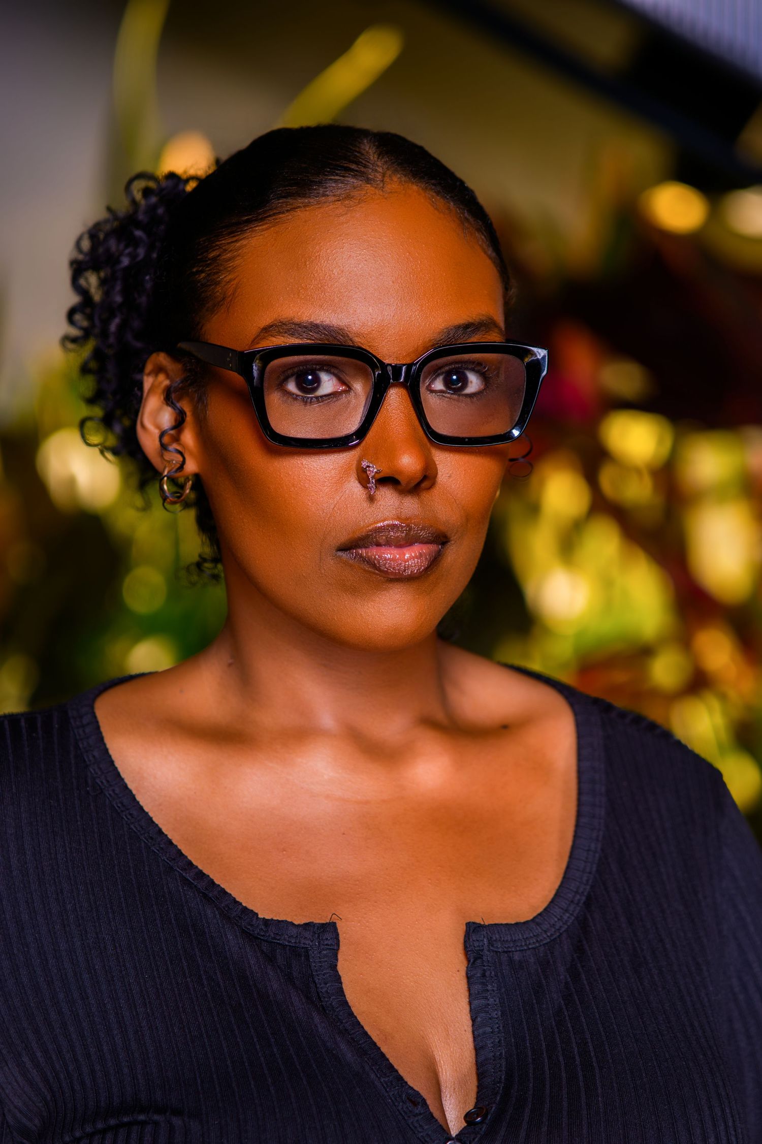 A young Black woman with black-framed glasses, hoop earrings, and a nose ring wearing a dark top.