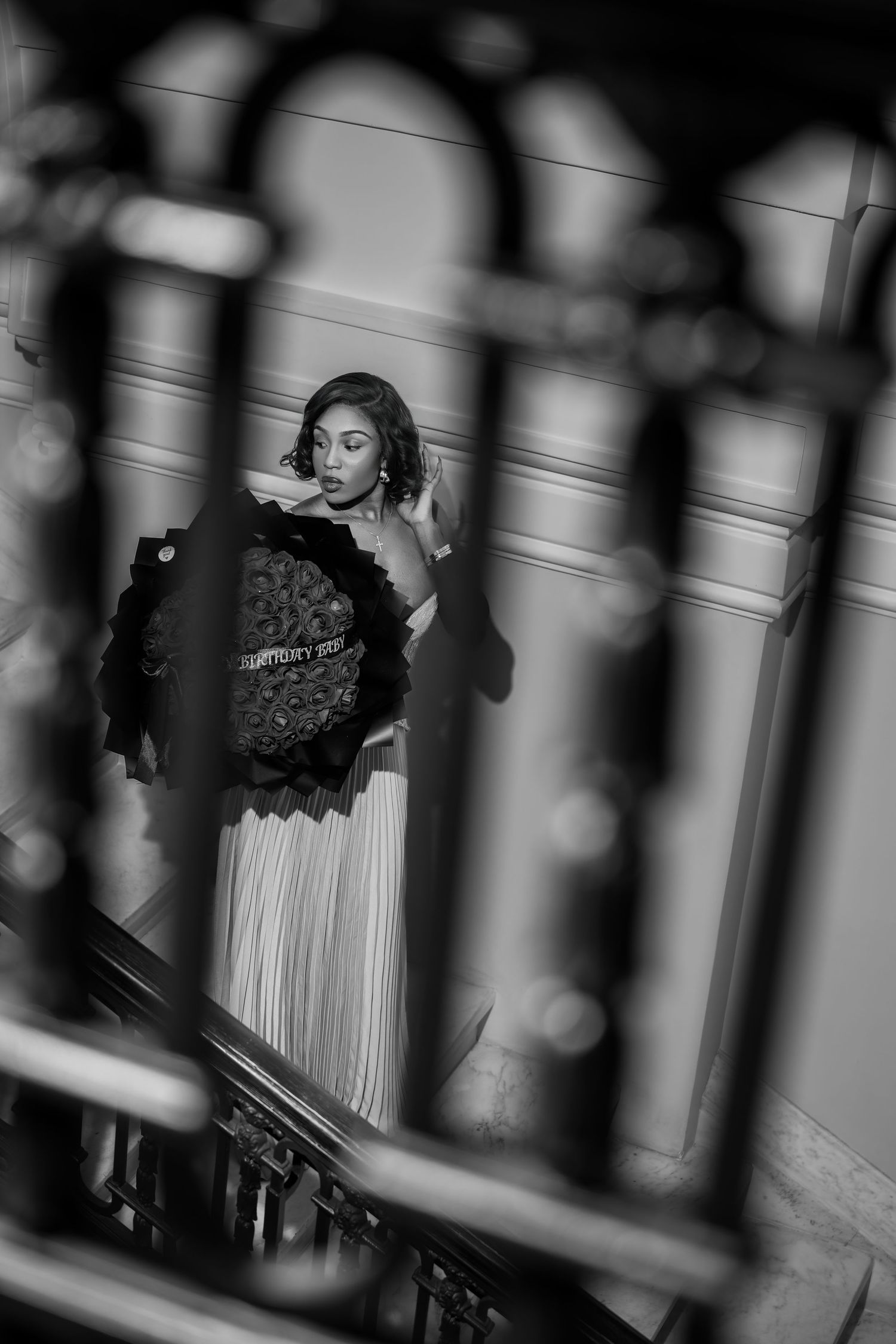 Black and white photo of elegant woman holding floral bouquet on grand staircase, viewed through ornate iron railing.