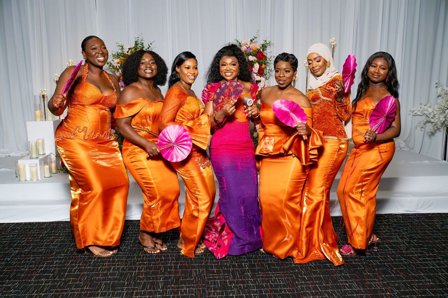 Wedding party members in vibrant orange and pink dresses pose together for a group photo.