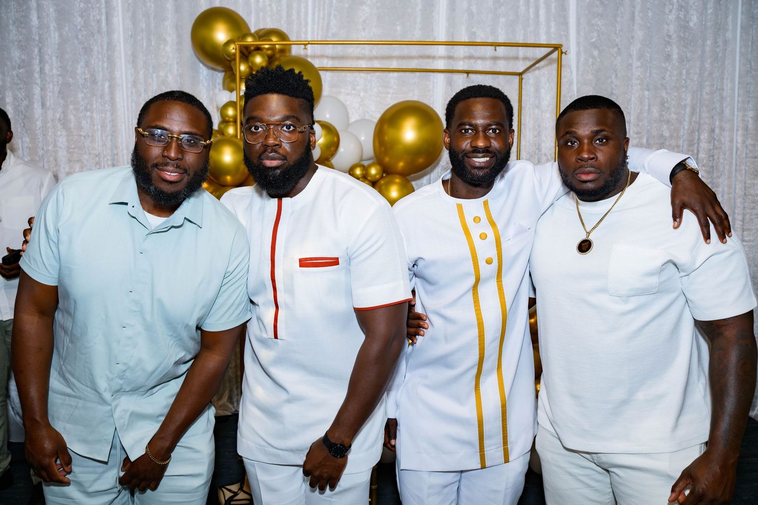 Four friends in casual white outfits pose together against a backdrop with gold balloons.