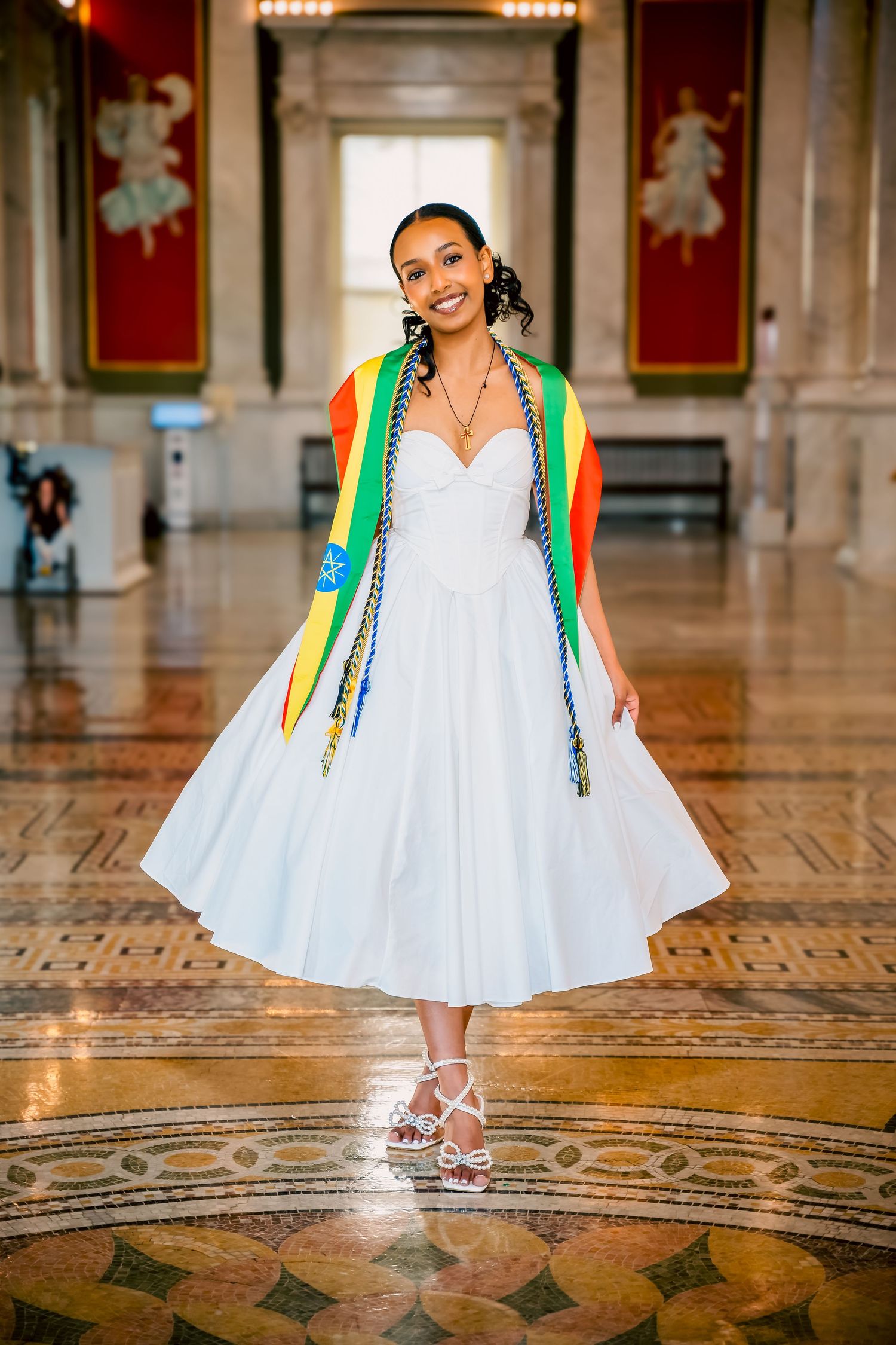 Woman in white dress with green and orange accents poses on ornate tiled floor in elegant hallway.