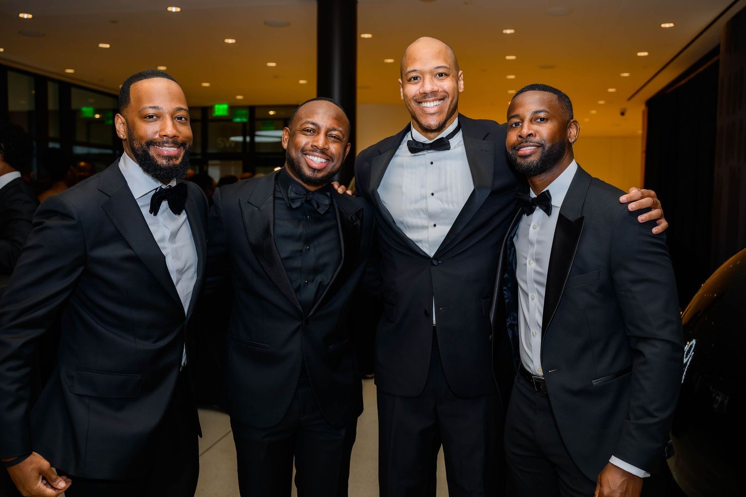 Four men in black tuxedos smiling together at a formal indoor event.