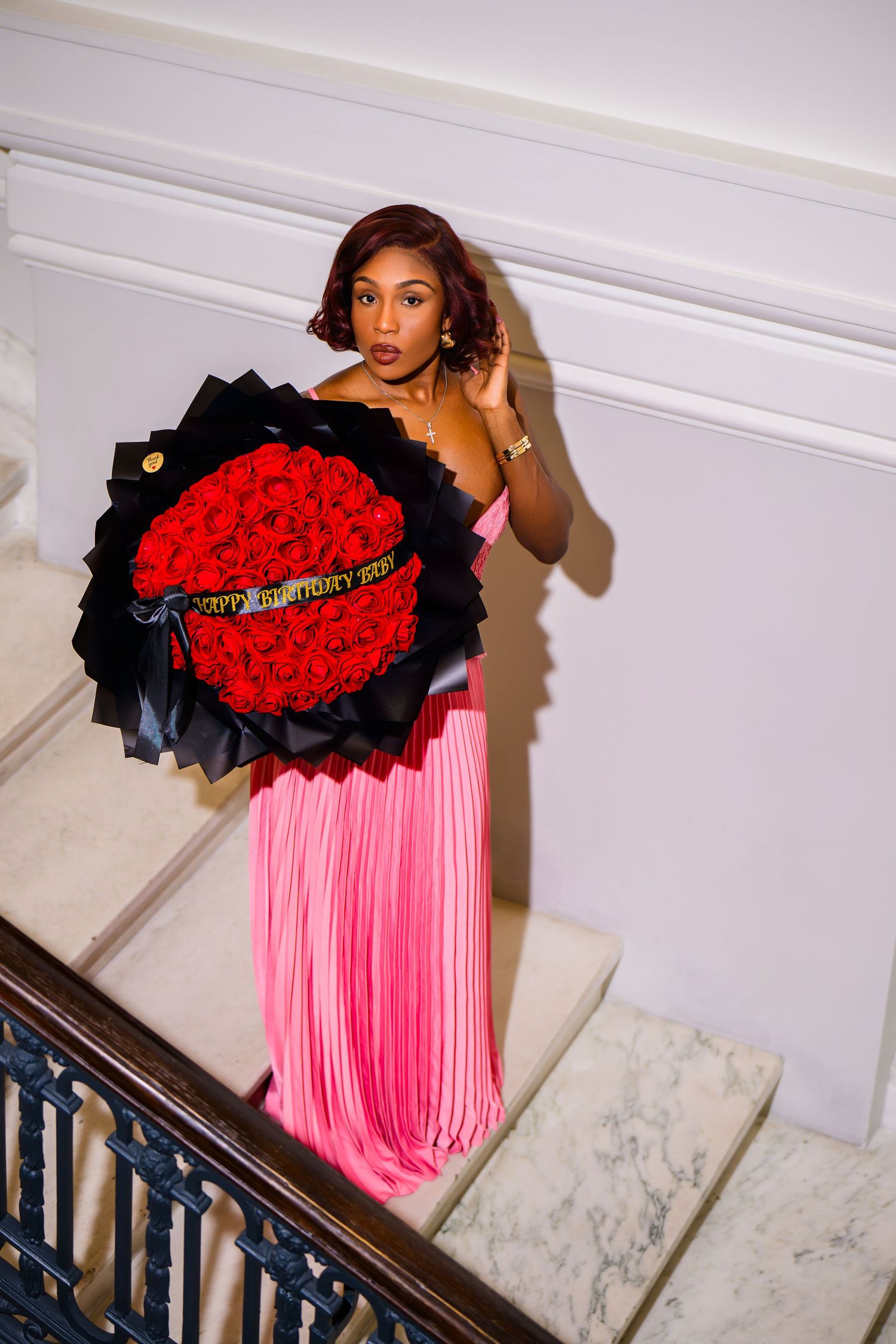Woman in pink fringe gown holding large red rose bouquet with Happy Birthday ribbon on marble staircase.