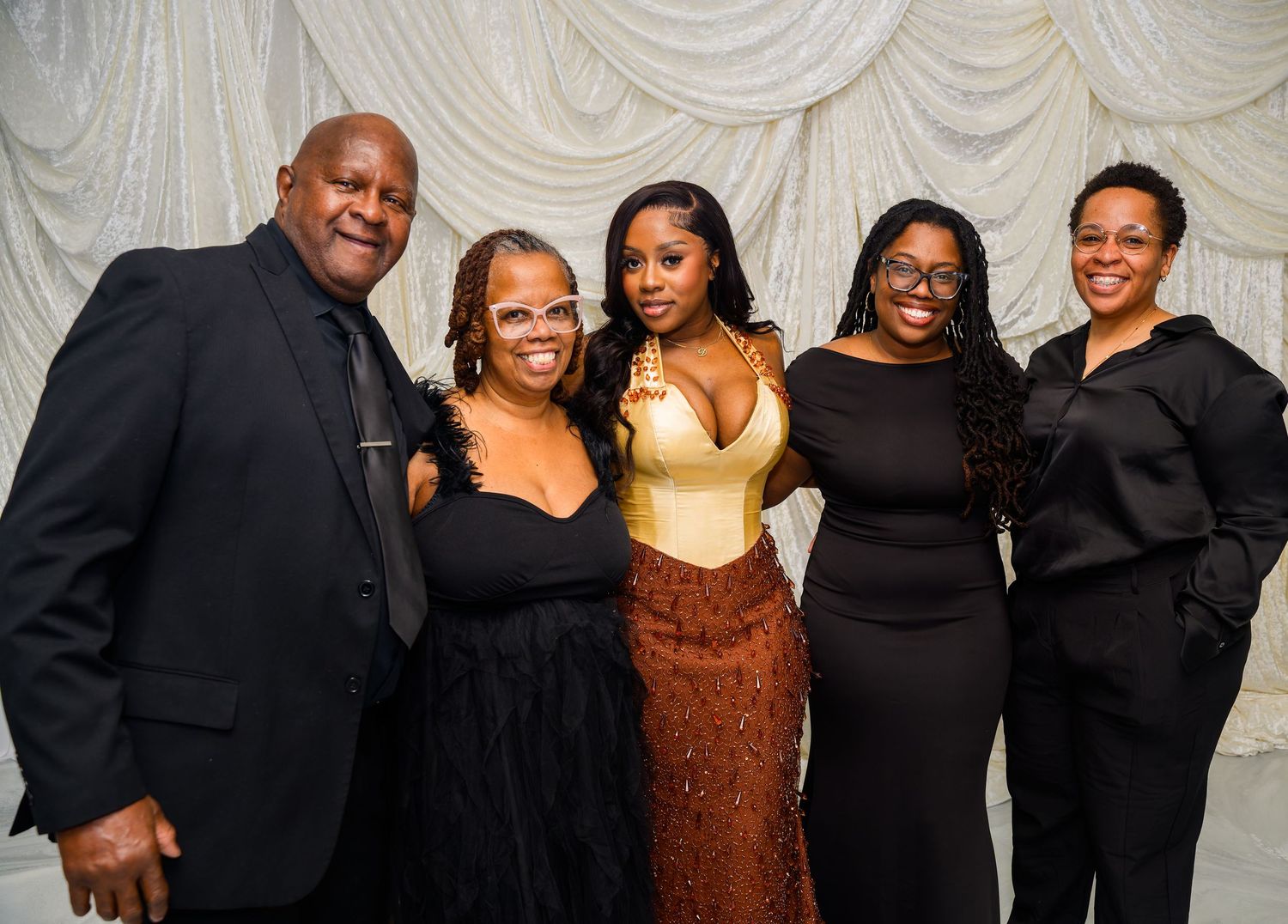 Five elegantly dressed people posing together at a formal event in front of a white draped backdrop.
