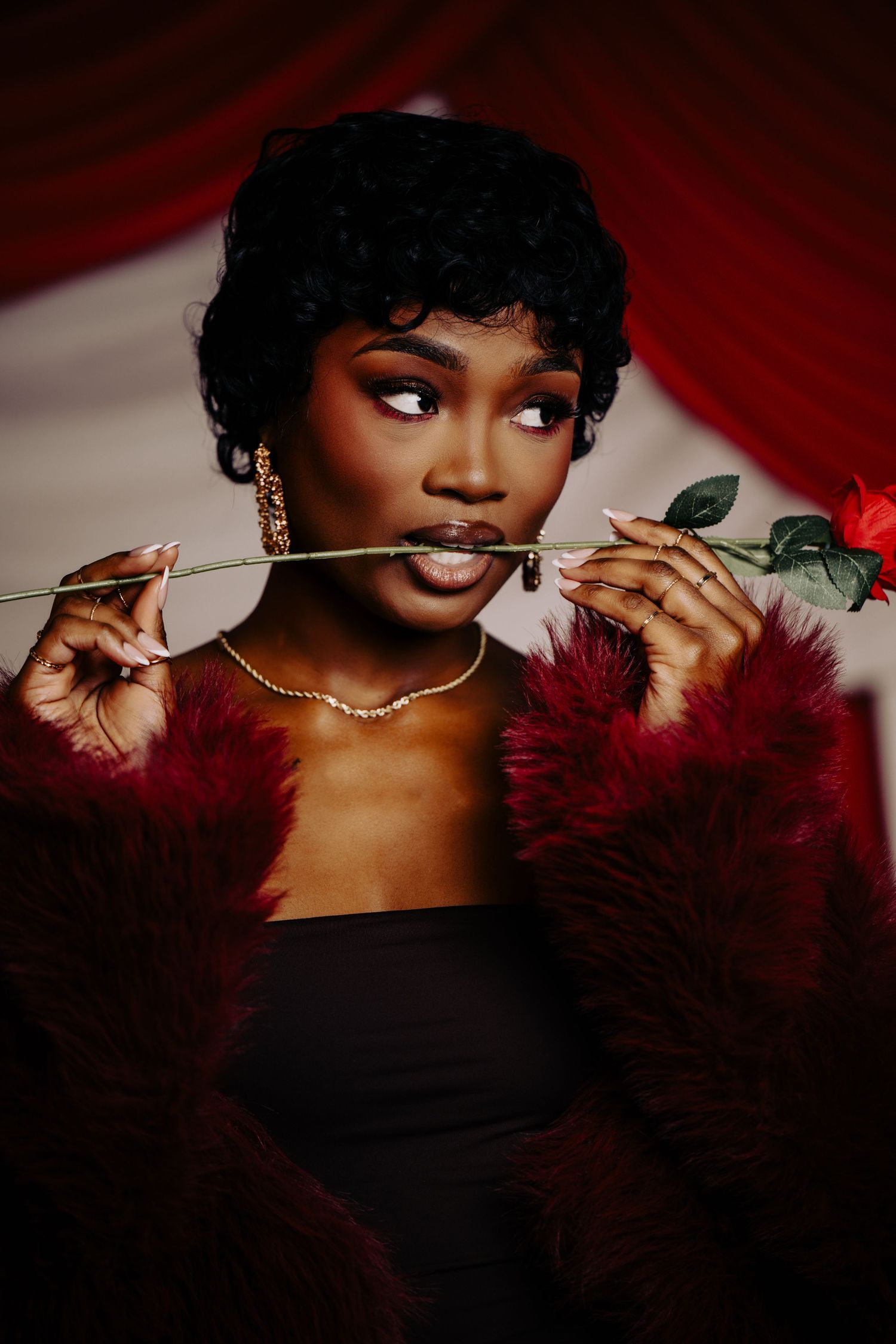 Glamorous portrait with sunglasses, red rose, burgundy fur stole and gold jewelry against dramatic red backdrop.