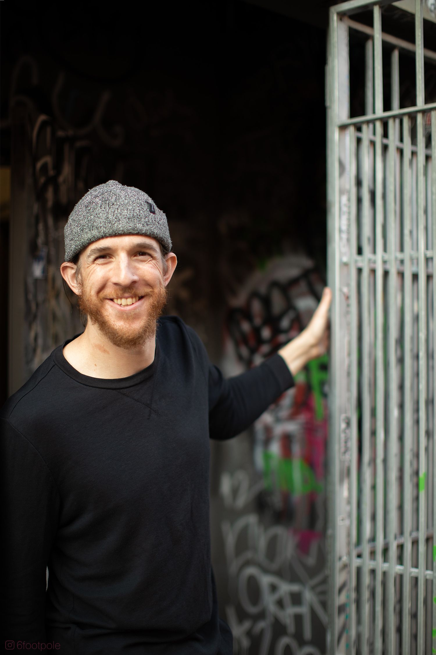A person wearing a black sweater and gray cap smiles against a graffiti wall background.