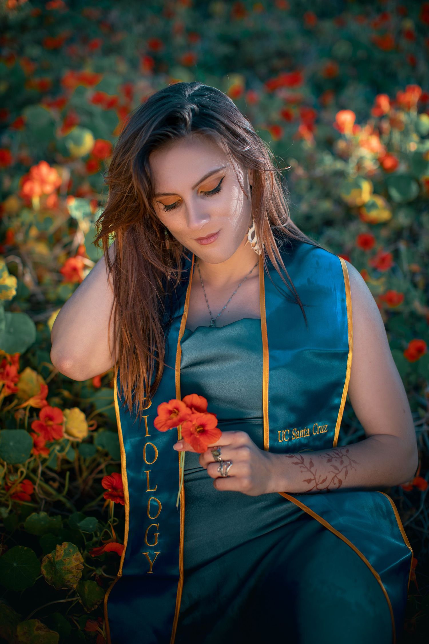 Graduate in academic stole poses among vibrant orange flowers in garden setting.