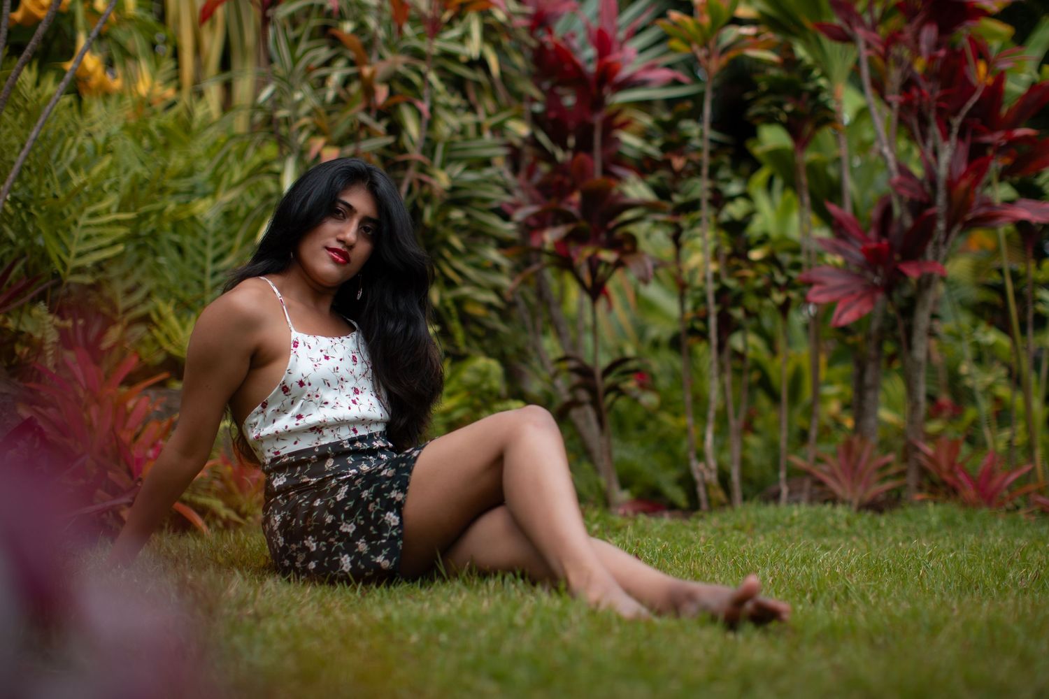 Model in white dress poses elegantly in a lush tropical garden setting with vibrant red foliage and green plants.