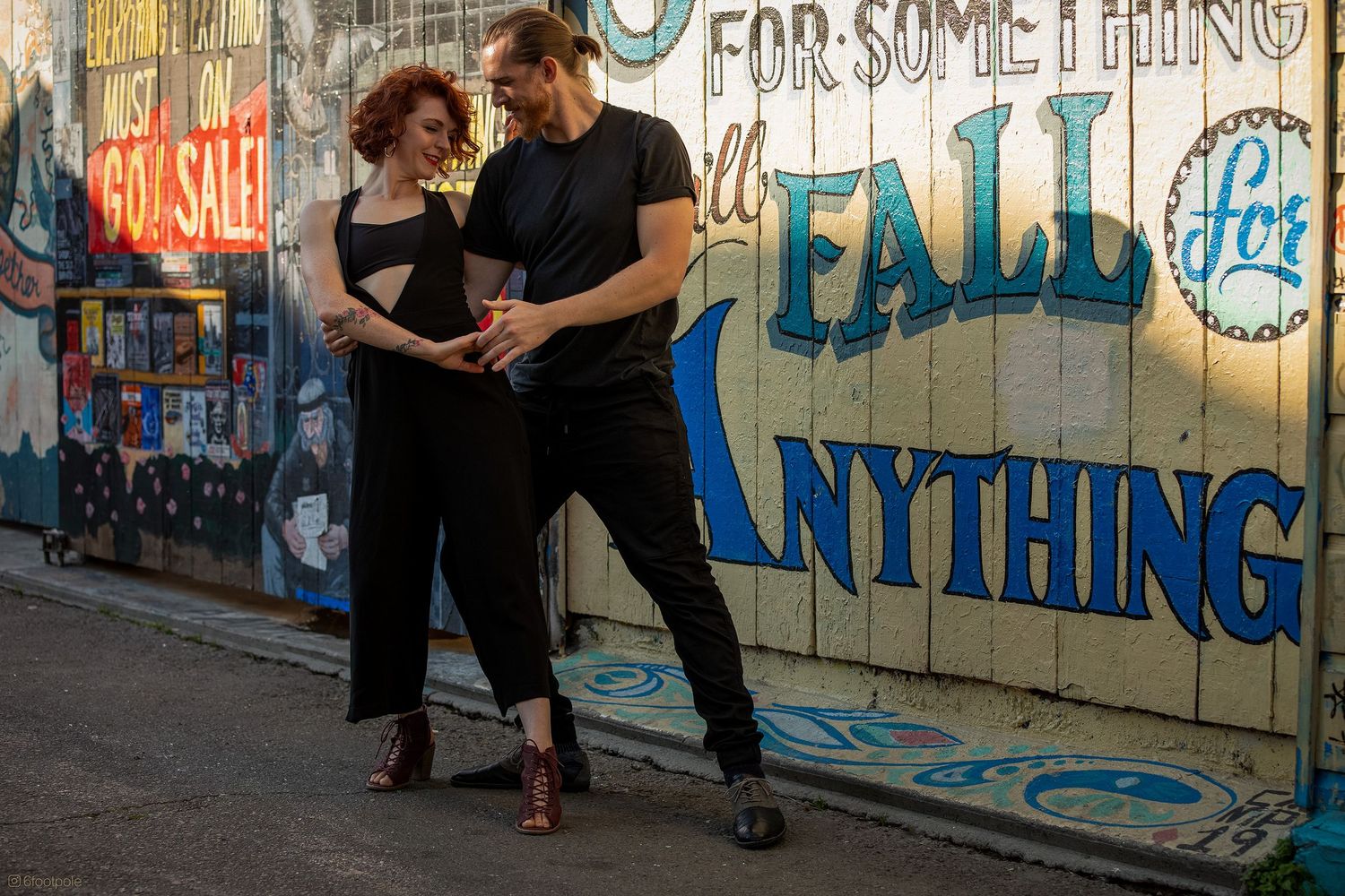 Two people dance together in front of a colorful graffiti wall with text reading 'For something' in an urban setting.