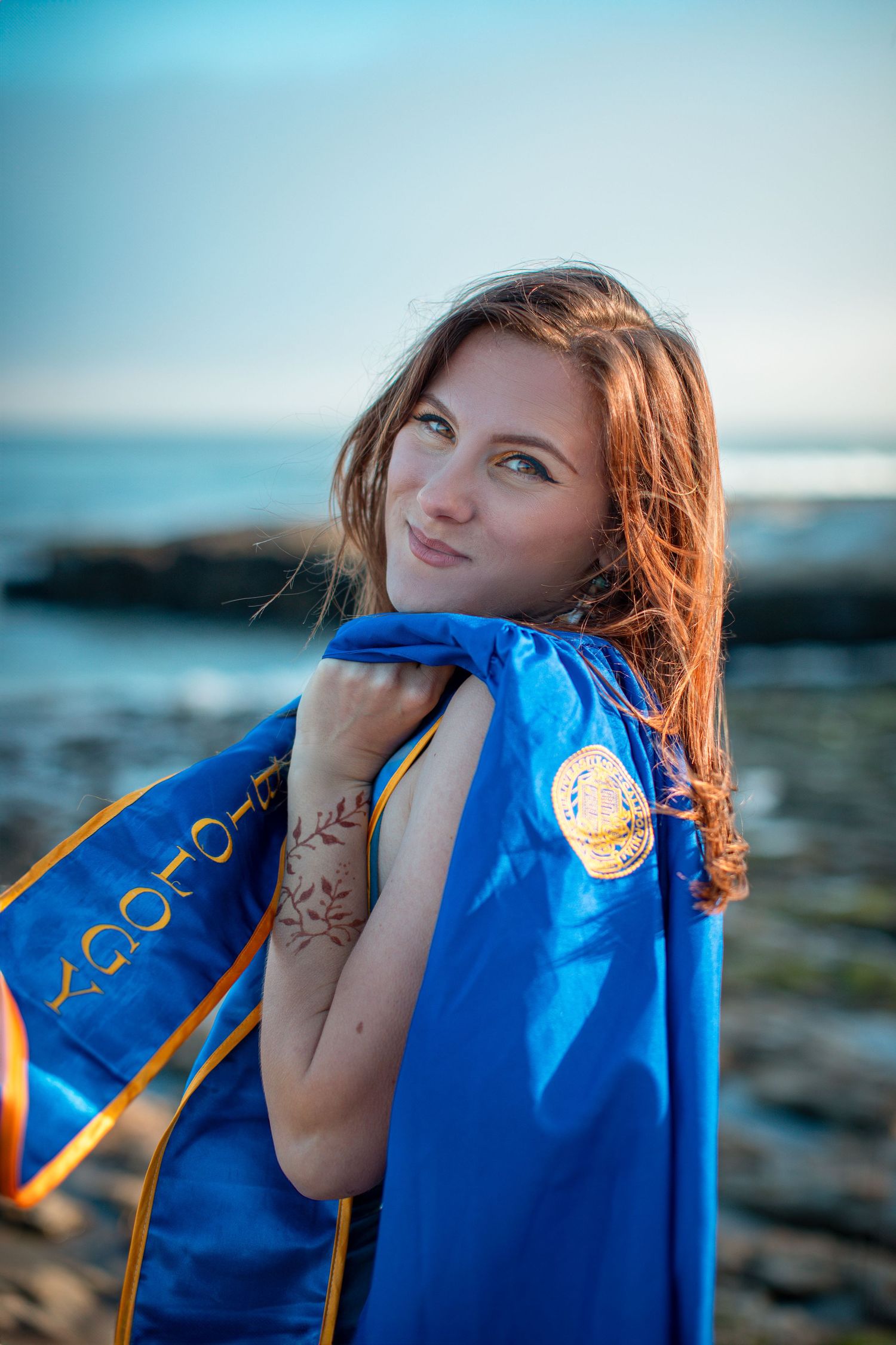 Graduate wrapped in blue graduation gown poses against coastal rocks and ocean backdrop.
