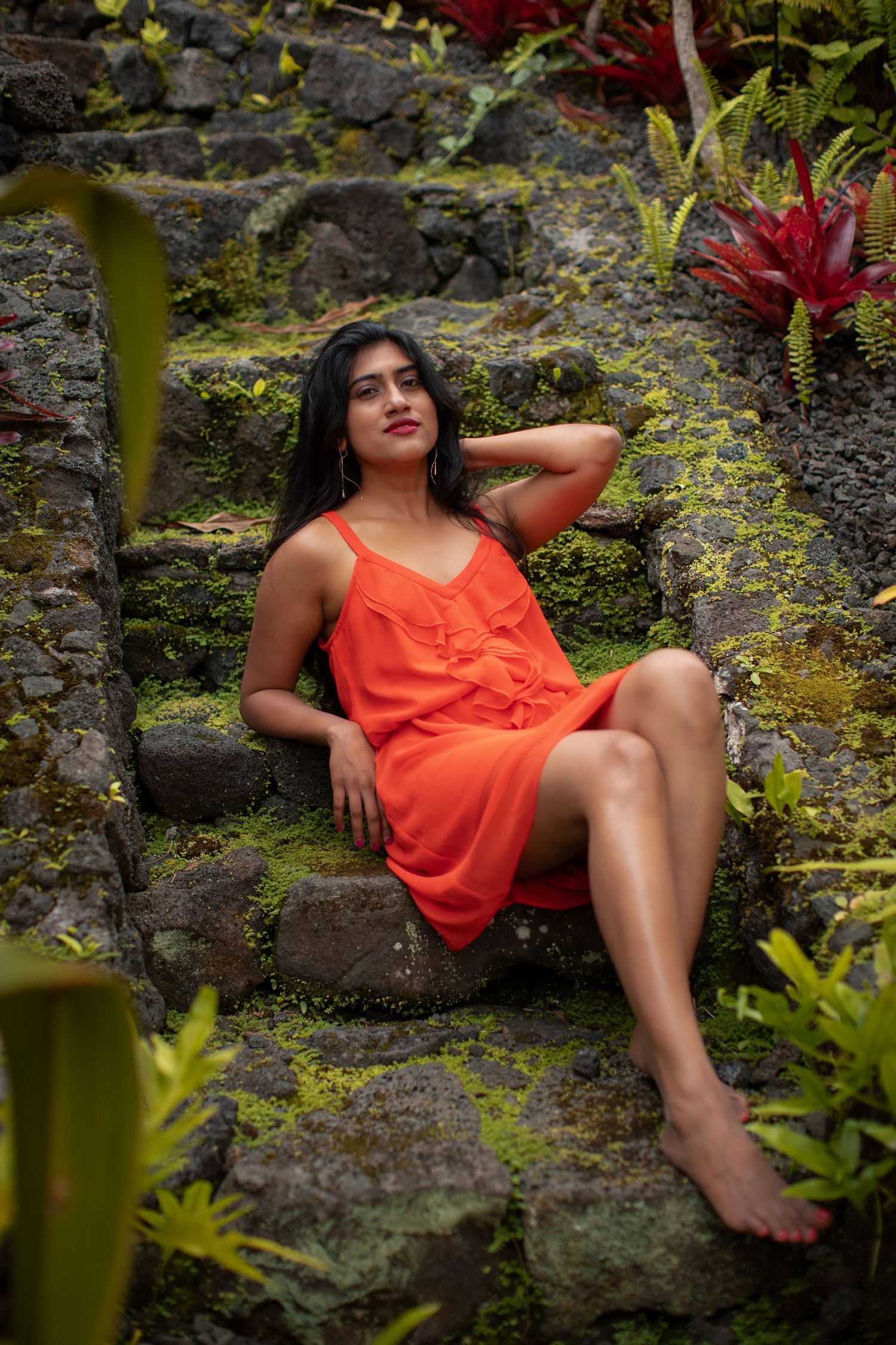 Model in coral dress poses elegantly on stone steps surrounded by tropical plants.