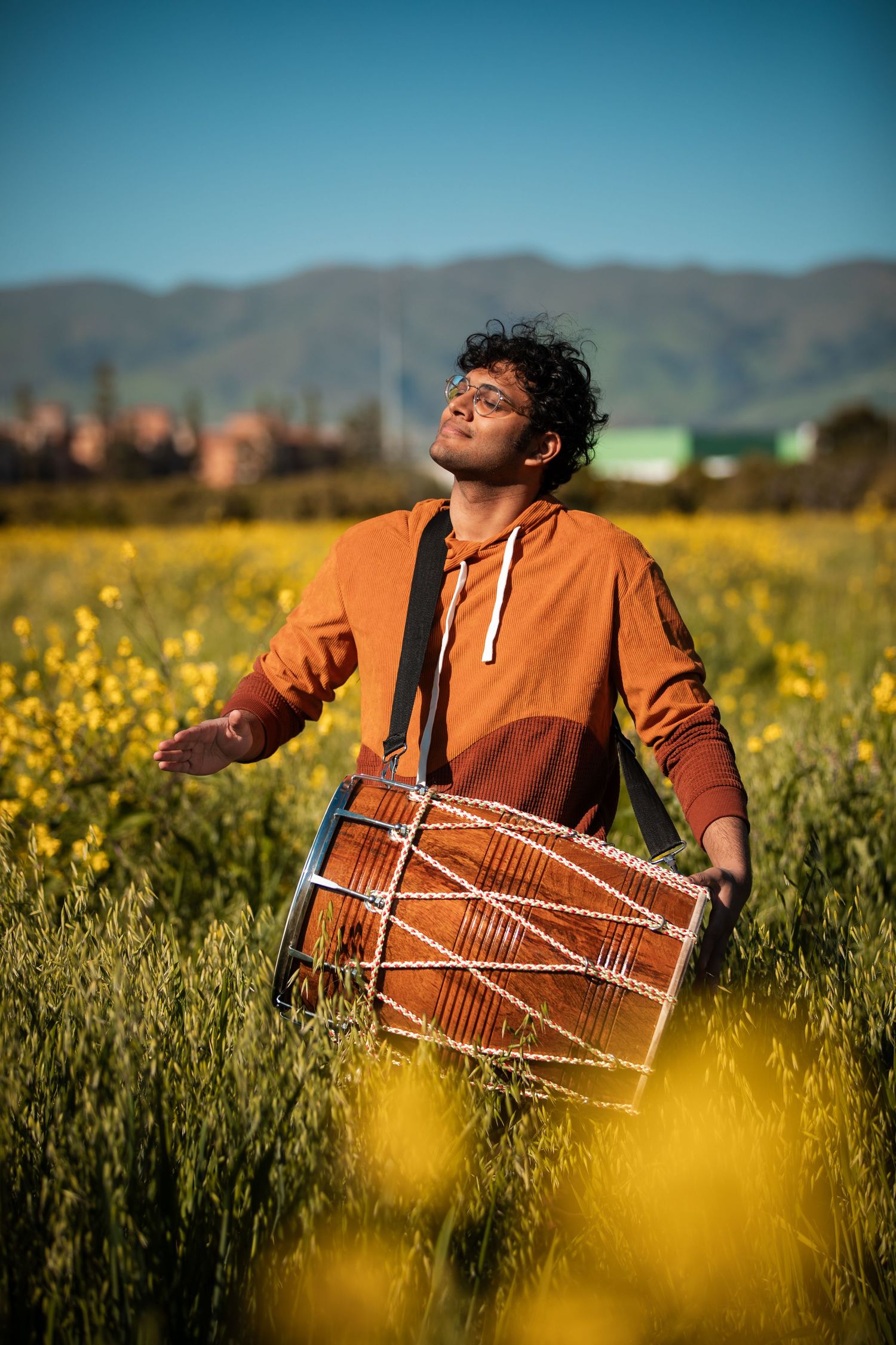 Musician playing dhol drum in orange hoodie amid yellow wildflower field with mountains.