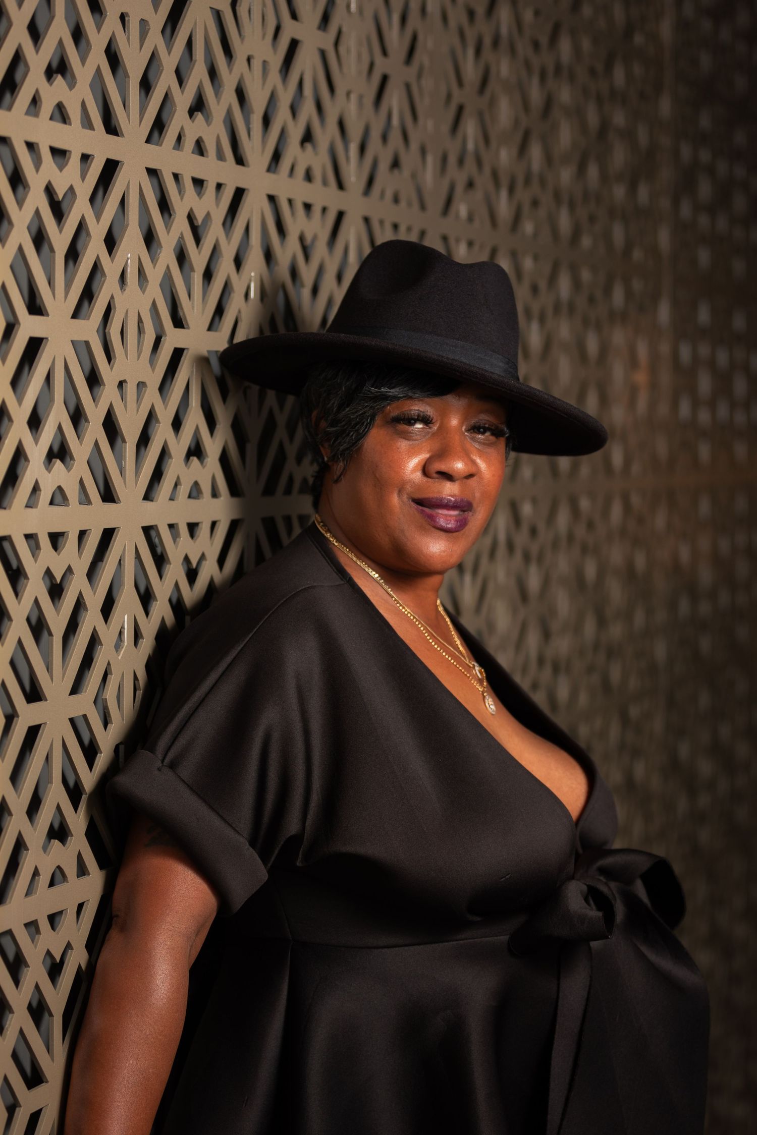 Dramatic portrait of someone in black dress and hat against textured geometric wall.