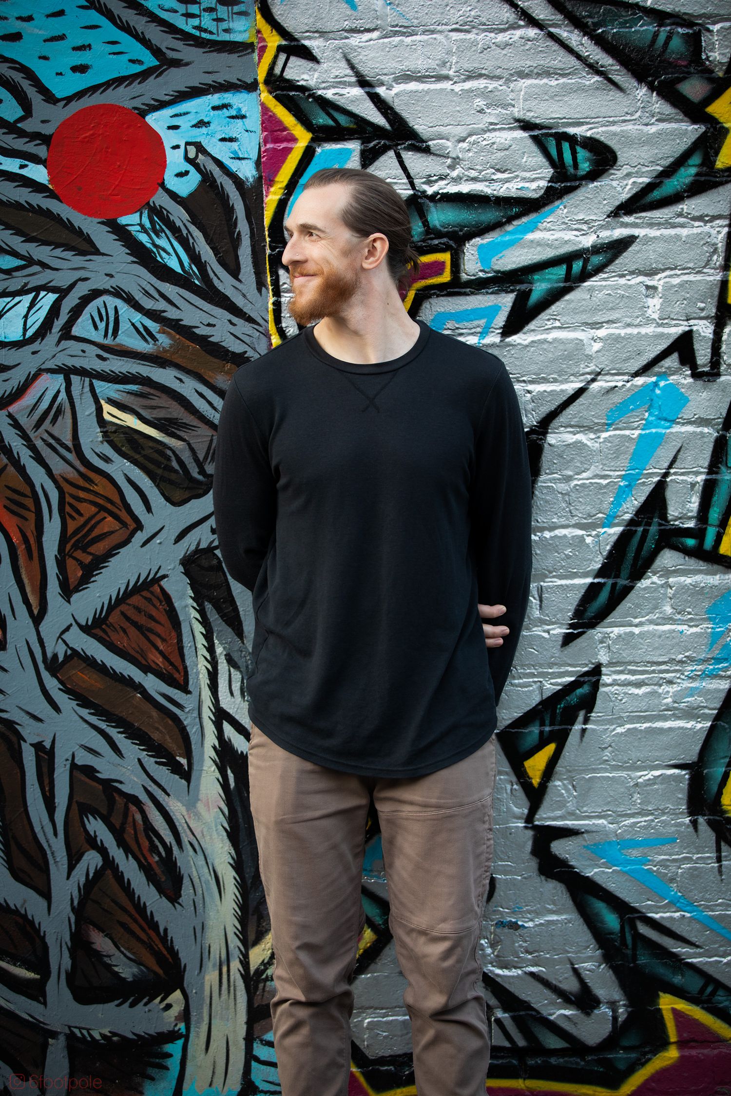 Person wearing black top and brown pants poses against graffiti wall in urban setting.