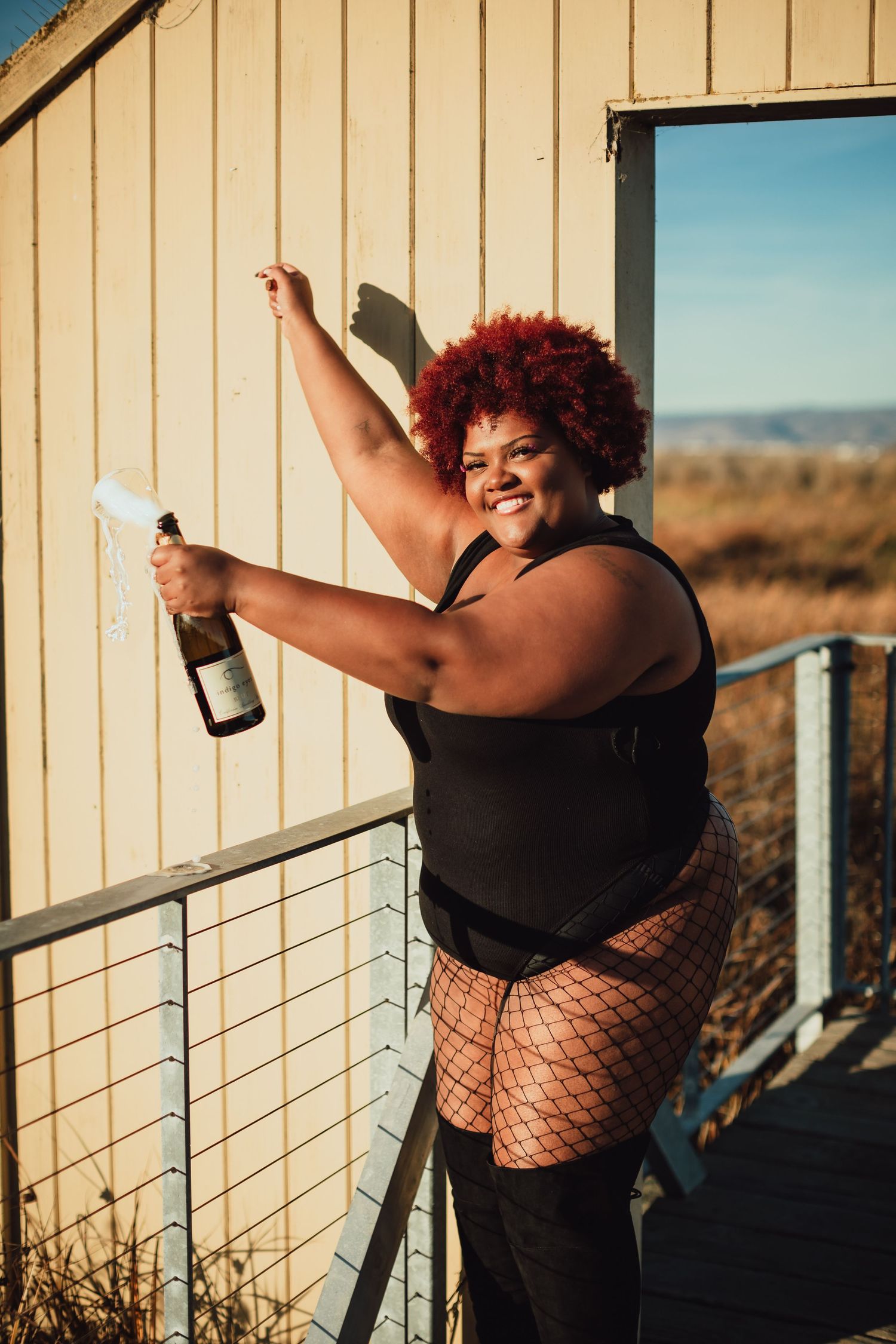 Woman in black outfit poses joyfully against wooden wall during outdoor photoshoot at sunset.