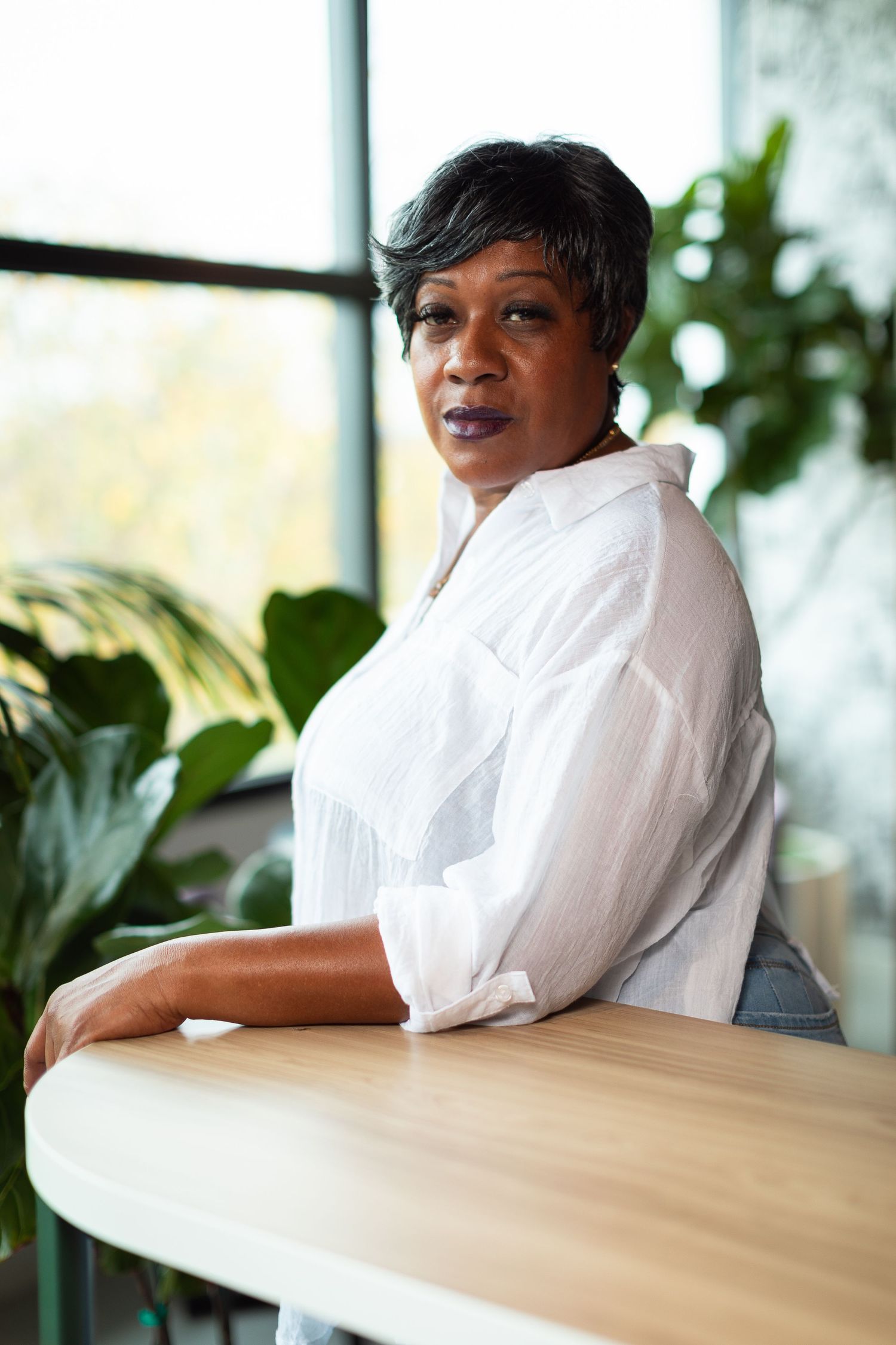 A person sits at a wooden table in a bright room with indoor plants.