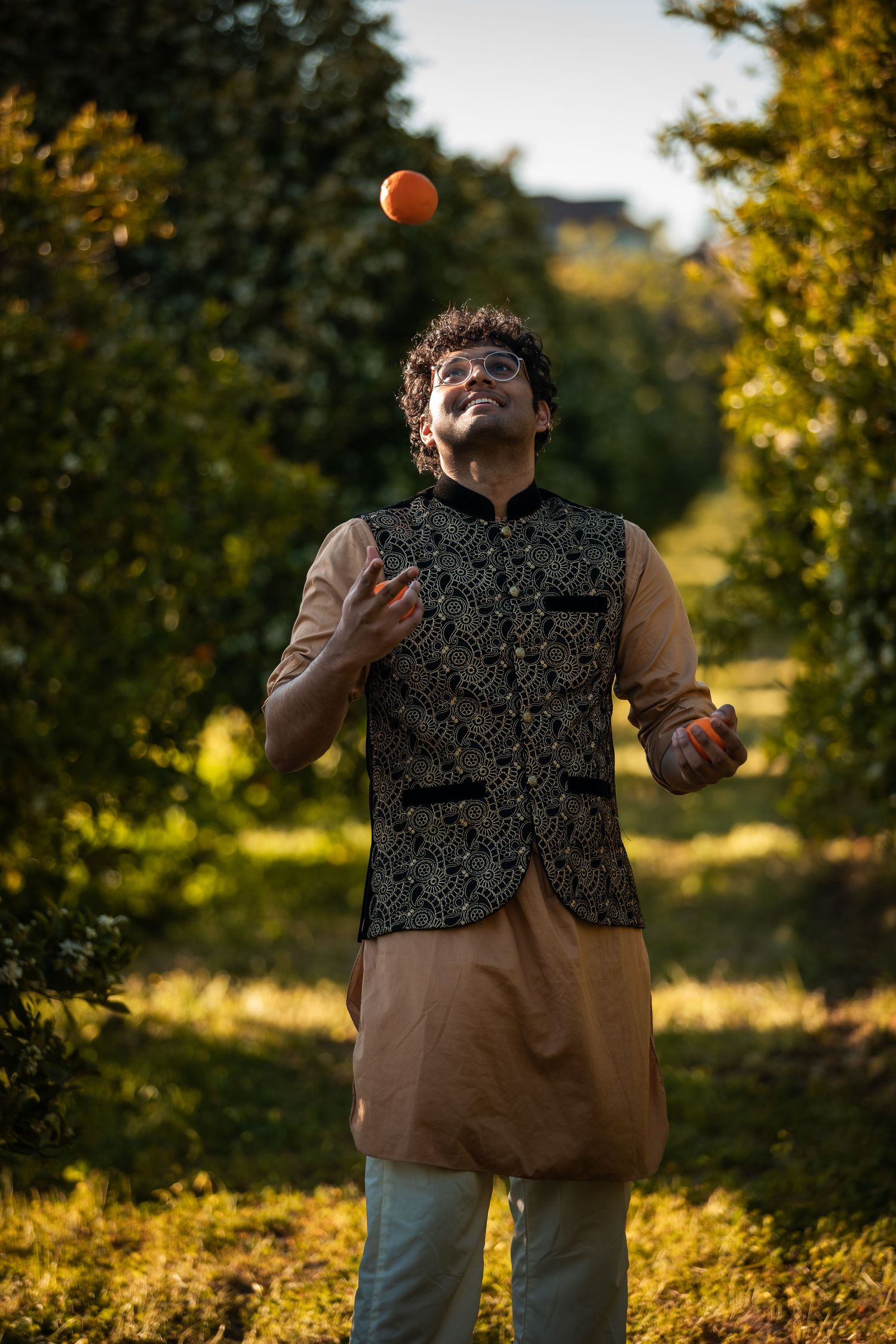 Person in patterned vest juggles orange in sunlit garden.