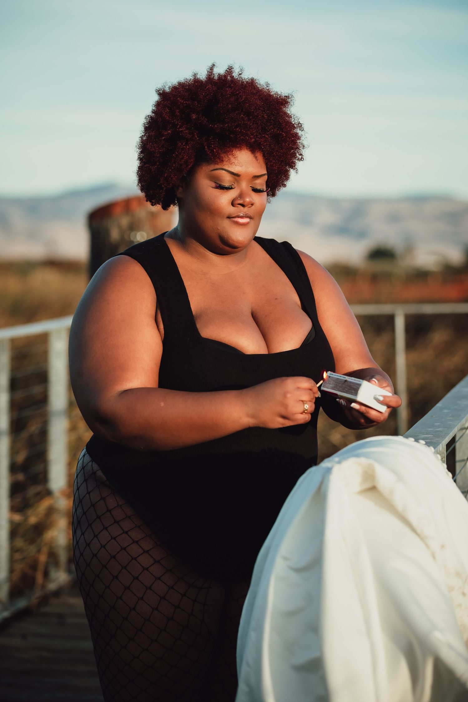 A person sits on a wooden deck examining a white dress during sunset.