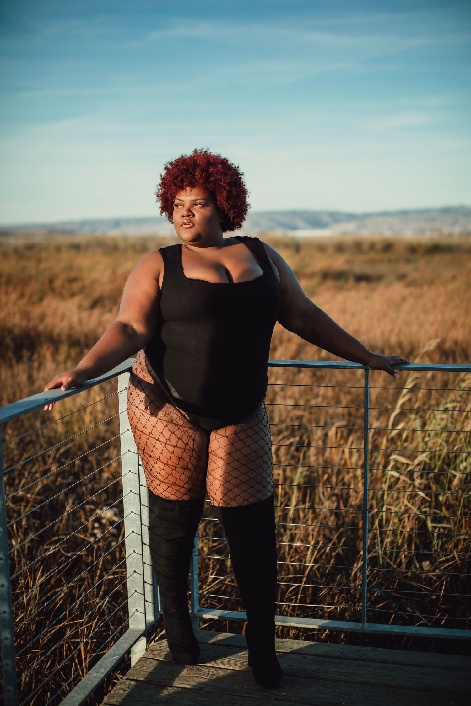 Artistic fashion portrait on wooden deck surrounded by tall grass at sunset.