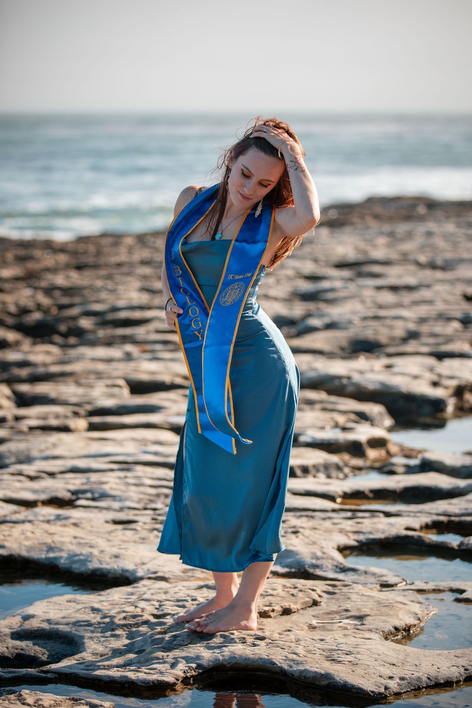 Graduate in blue dress poses gracefully on rocky coastal shoreline with ocean waves in background.