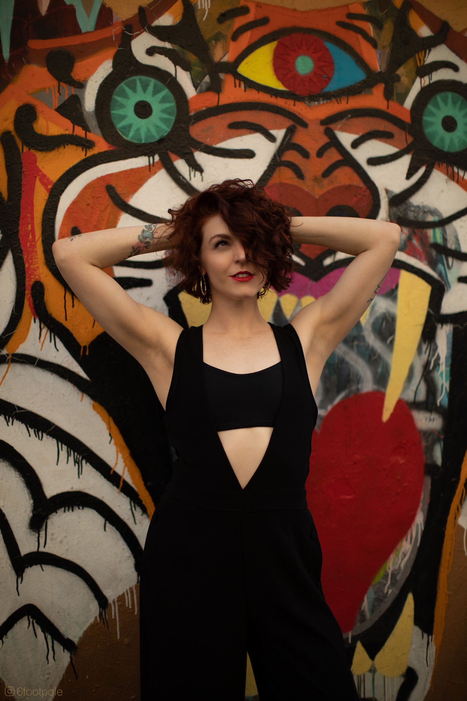 Person in black dress poses playfully against vibrant street art mural with circular eye designs and bold colors.
