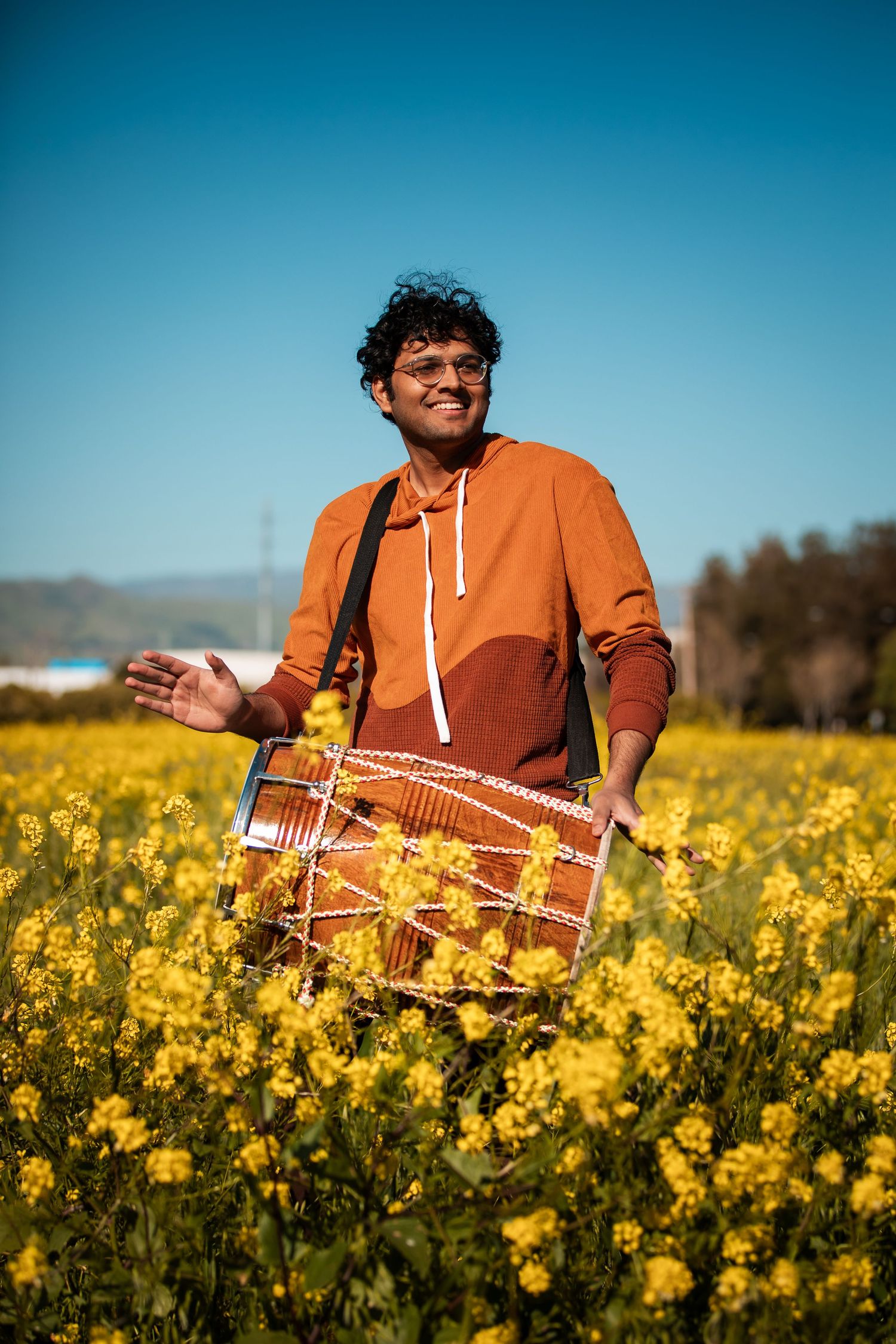 Person in orange sweater sits among vibrant yellow canola flowers under blue sky.