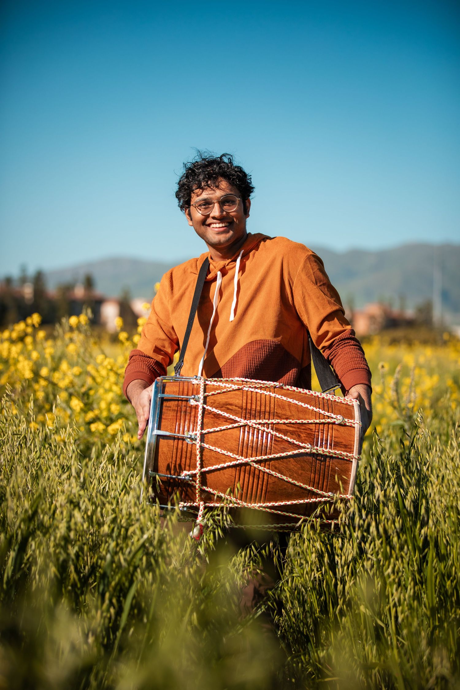 Joyful drummer playing traditional dhol in mustard field against blue sky.