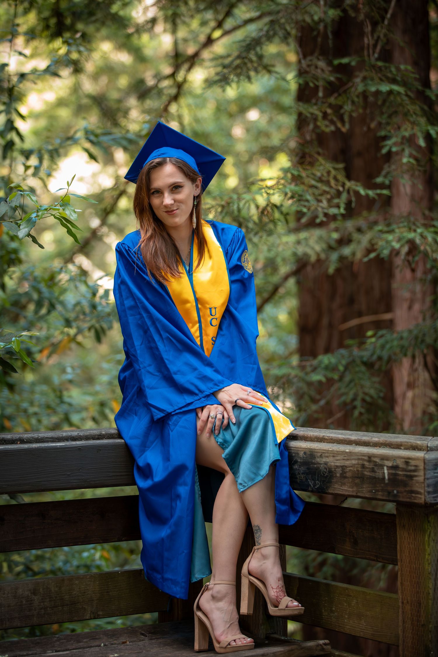 Graduation photos in blue cap and gown on wooden bridge in forest setting.