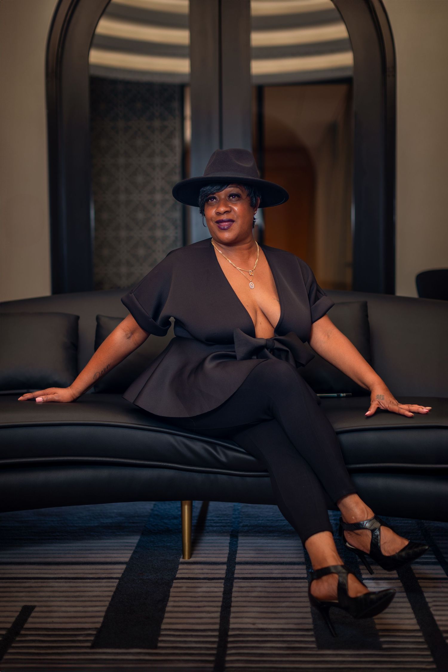 Stylish figure in black outfit and wide-brimmed hat poses dramatically on dark leather couch.