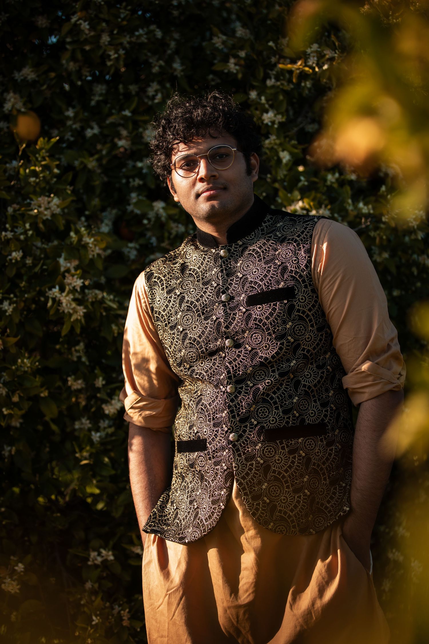 Figure in textured vest poses against garden backdrop in warm lighting.