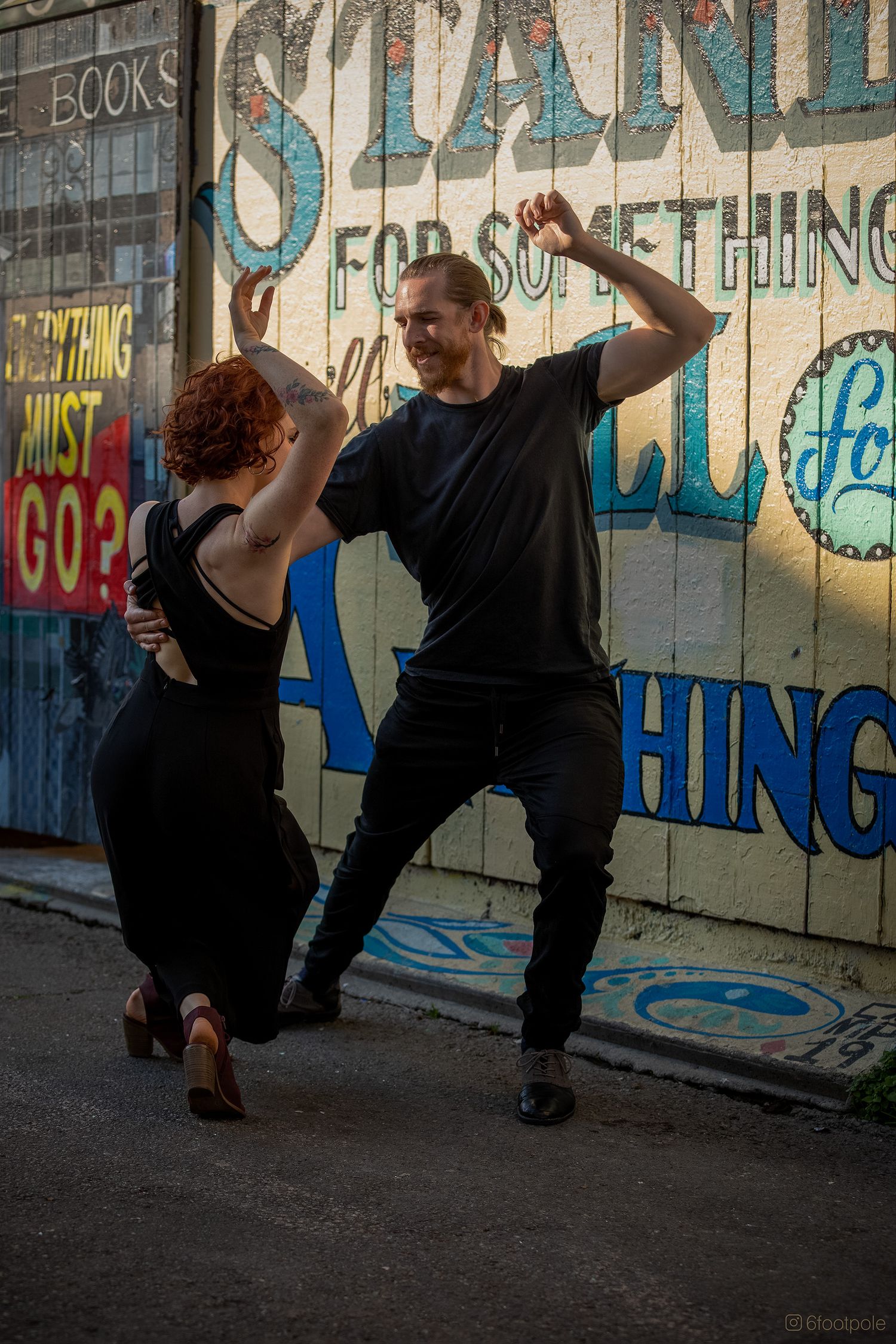 Two dancers perform a contemporary dance sequence against a graffiti wall in black clothing.