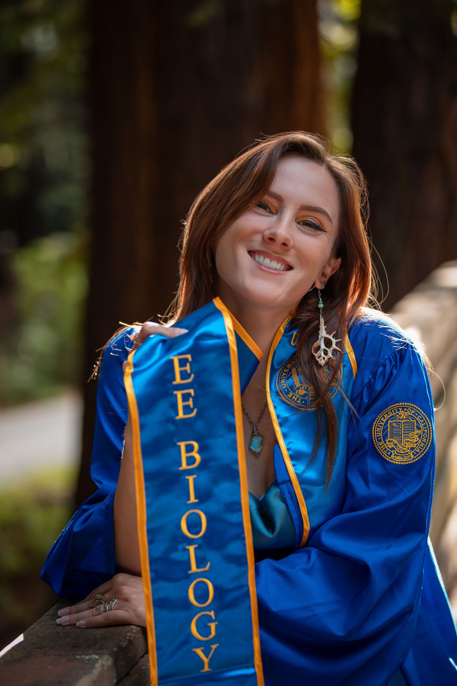 Marine biology graduate wearing blue and gold academic stole smiles during outdoor graduation photoshoot.