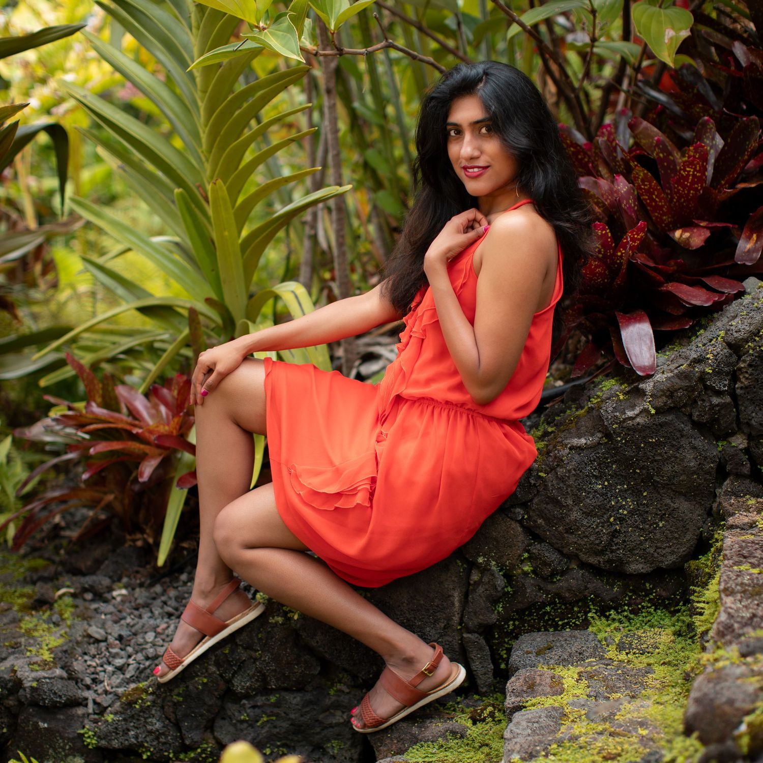Woman in coral sundress sits on stone wall surrounded by tropical foliage in garden setting.